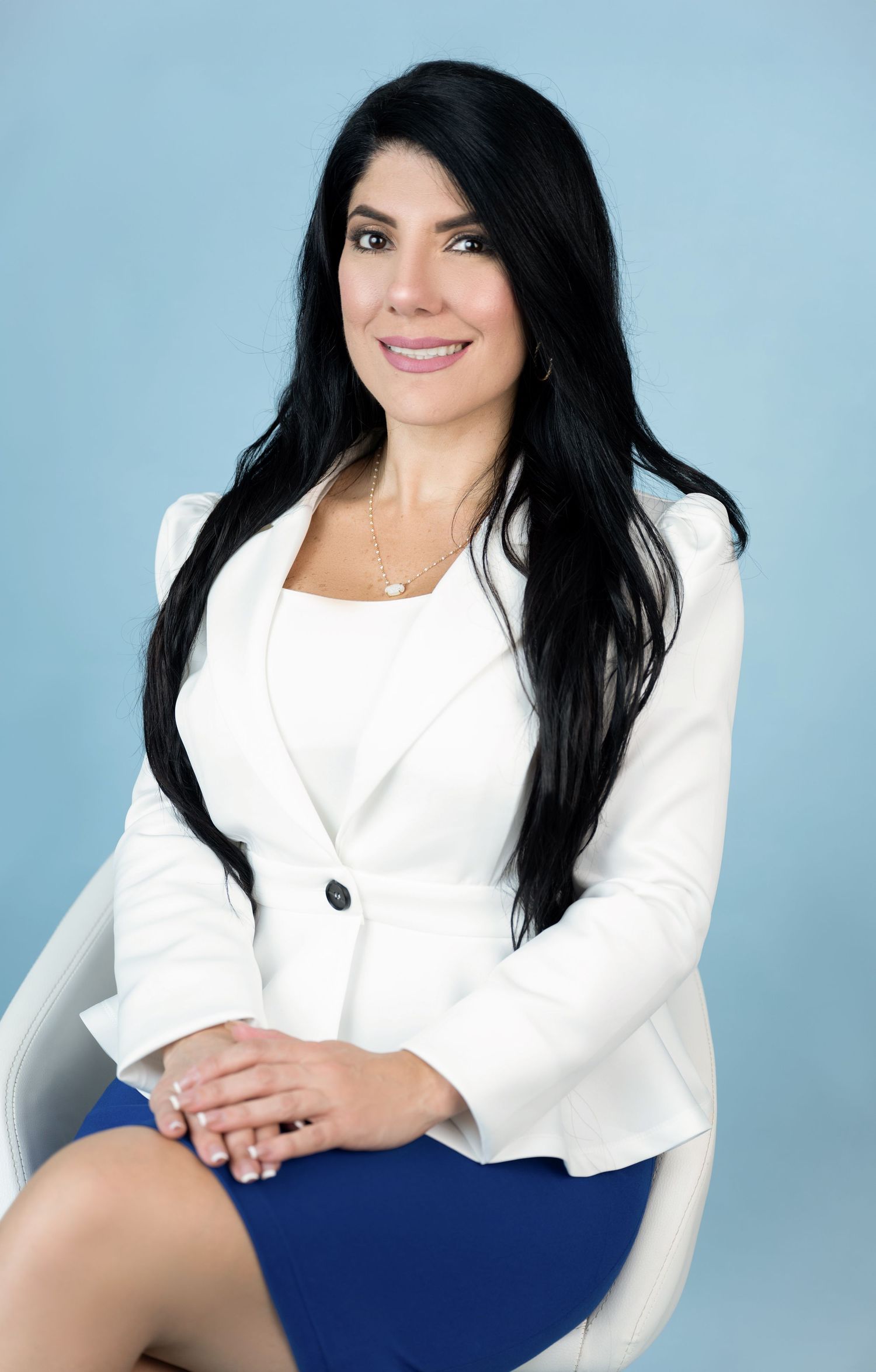 Smiling woman with long black hair in a white blazer and blue skirt sits with hands folded against a light blue studio background.