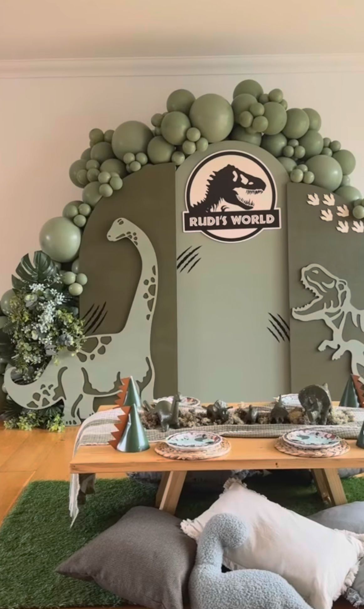 Dinosaur-themed party setup with sage green balloon arch, wooden table, and prehistoric wall decor on artificial grass floor.