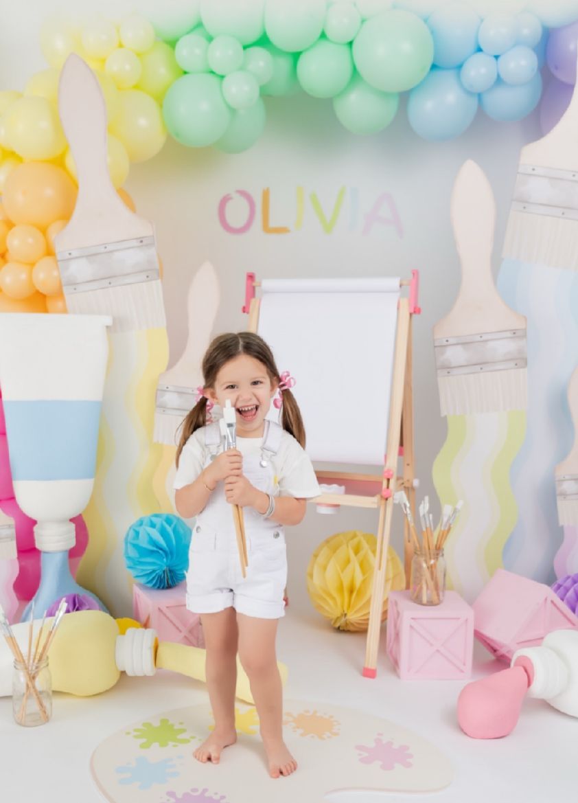 Colorful pastel balloon arch and party decorations create a festive backdrop for a child's celebration with white and pink accents.