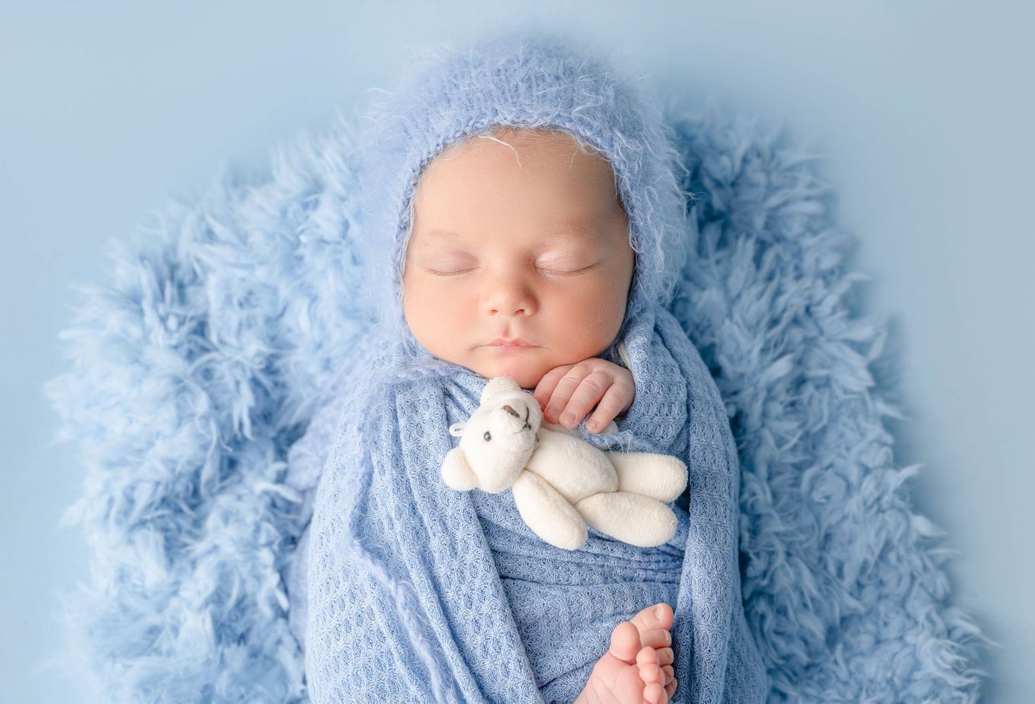 Sleeping newborn baby wrapped in blue knit blanket wearing bonnet, holding white teddy bear on fluffy blue background.