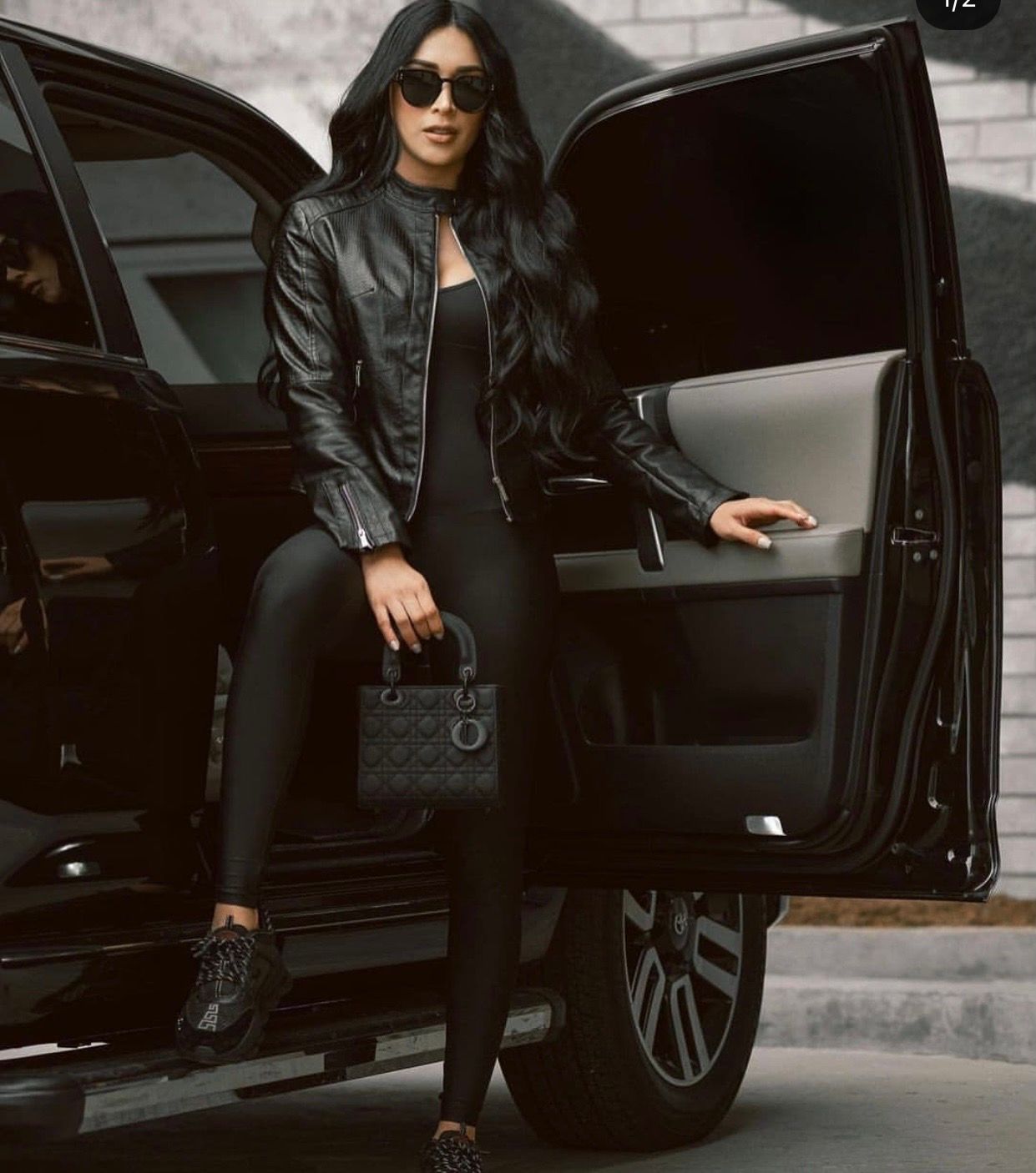 Woman in all-black outfit with leather jacket and sunglasses sitting in luxury SUV doorway holding a Dior handbag.
