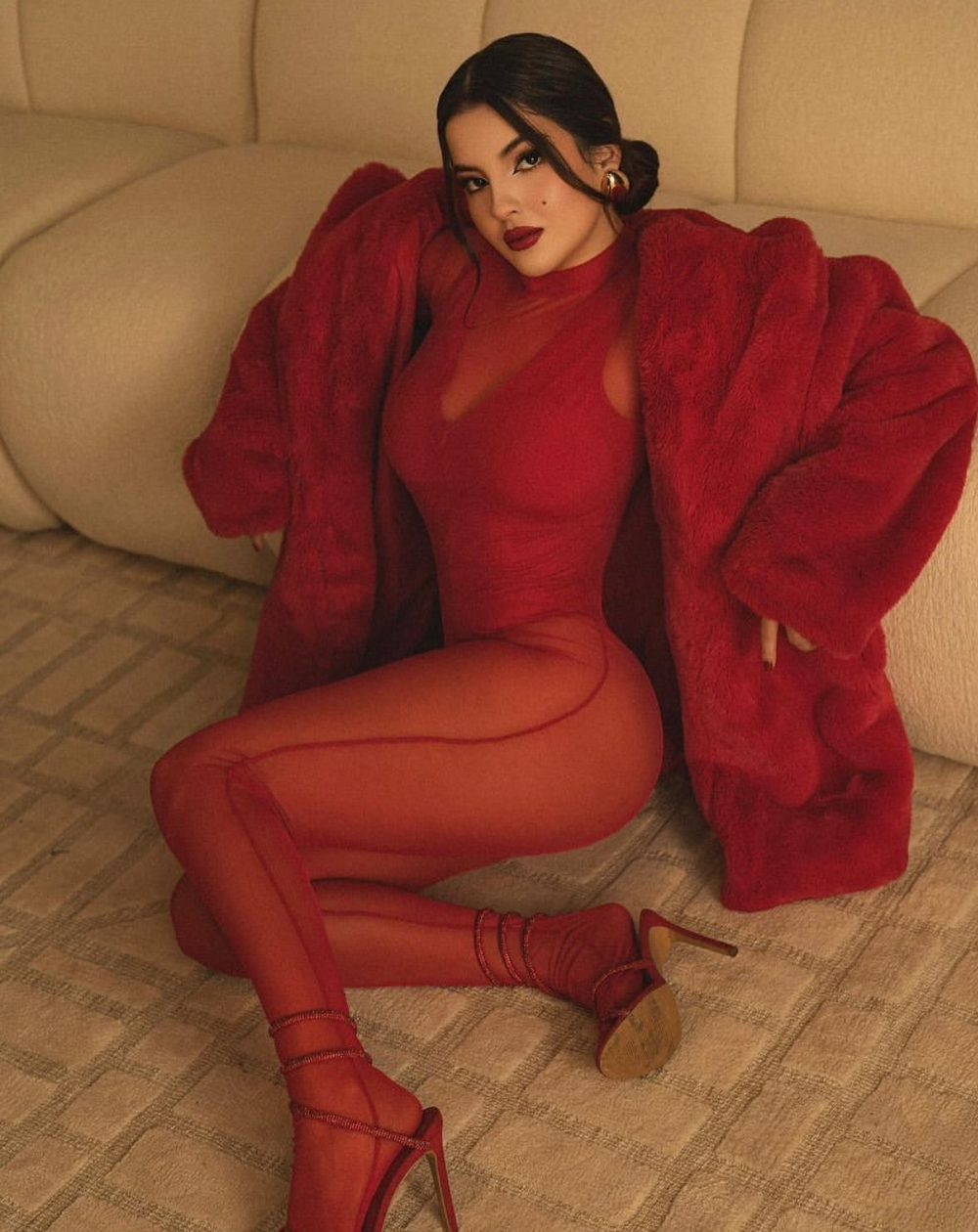 Woman in all-red outfit with faux fur coat, bodysuit, leggings, and heels, posing elegantly on a beige carpet.