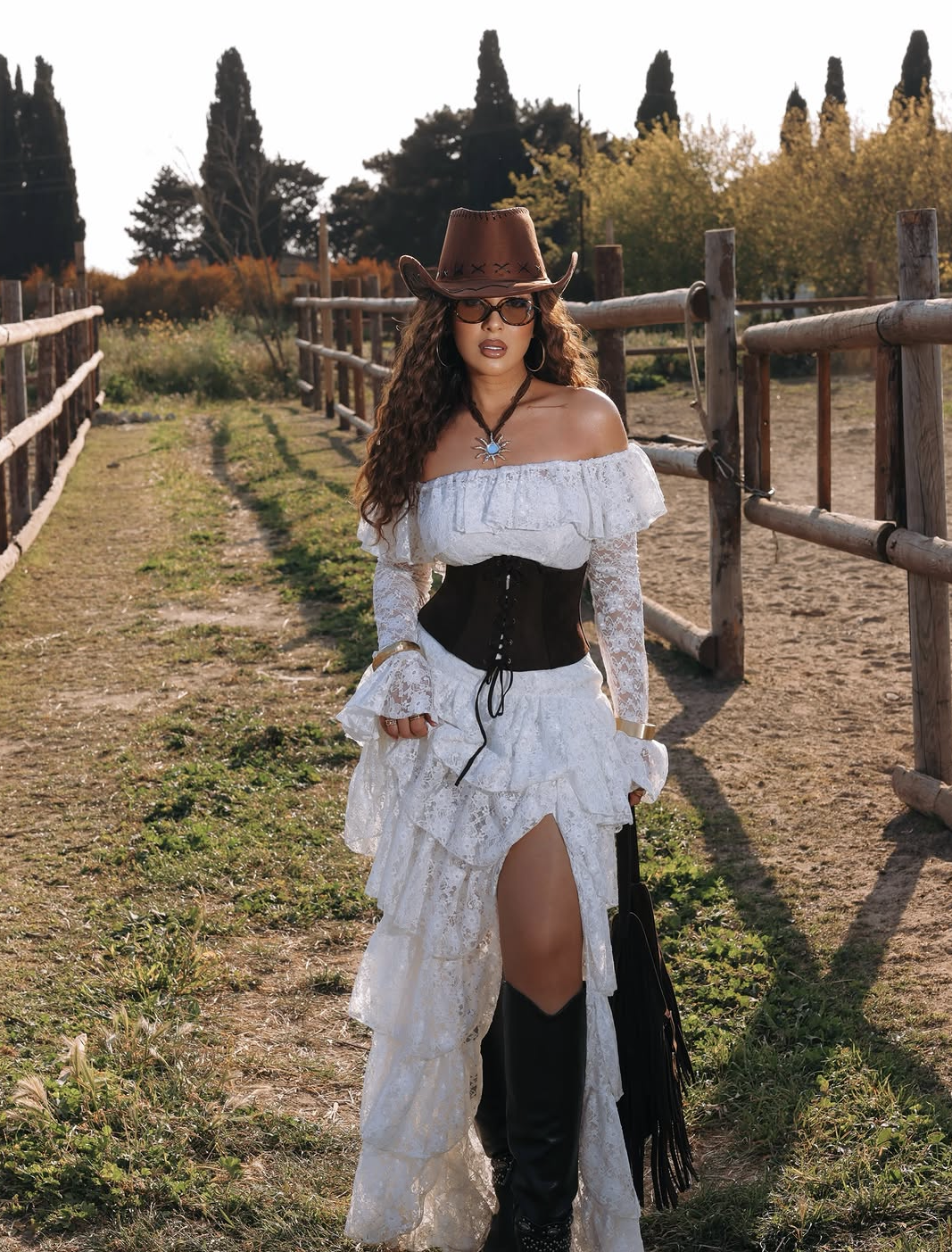 Woman in white lace off-shoulder dress, corset belt, brown cowboy hat, and black fringe boots on ranch path.