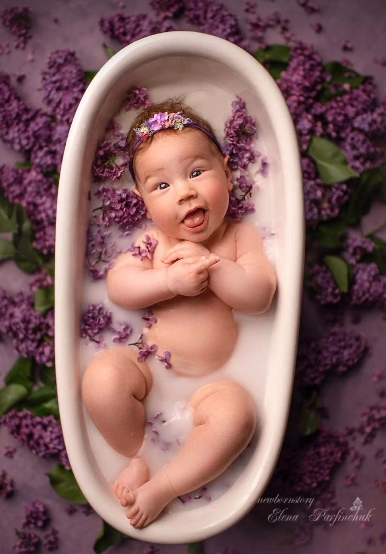 Smiling newborn baby in a white mini bathtub surrounded by purple lilac flowers, wearing a floral headband.