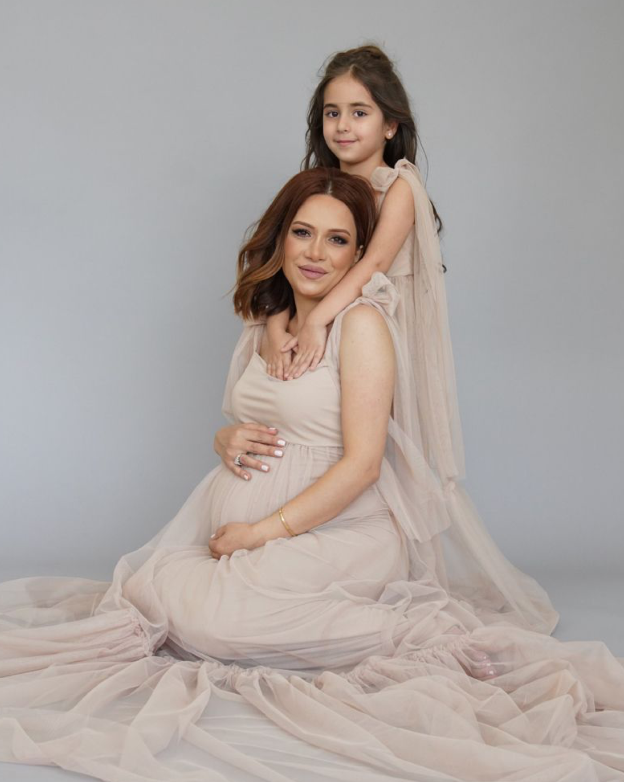 Pregnant woman in blush tulle gown sits with young daughter draped over her shoulders in a studio maternity portrait.