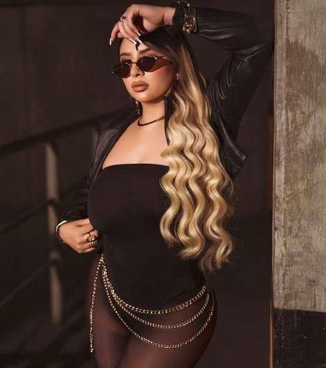 Woman in black outfit with chain belt, blonde wavy hair, sunglasses, and leather jacket poses confidently.