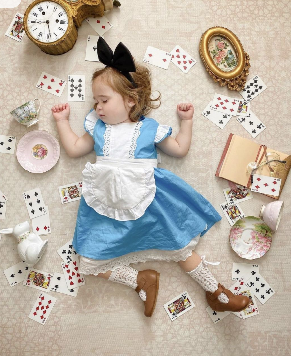 Toddler dressed as Alice in Wonderland sleeping surrounded by playing cards, a clock, teacups, and books.