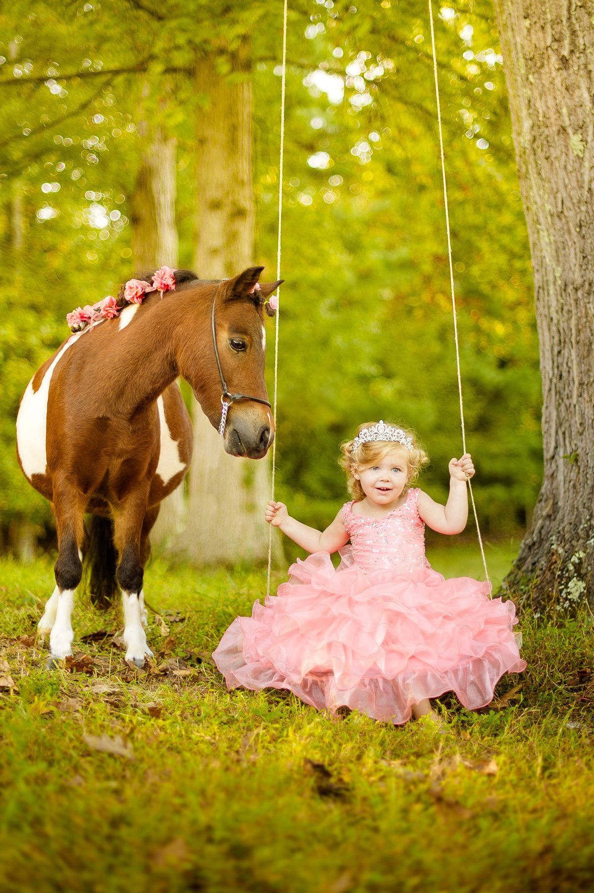 A little girl in a pink princess dress and tiara sits on a rope swing beside a mini horse in a lush green park.