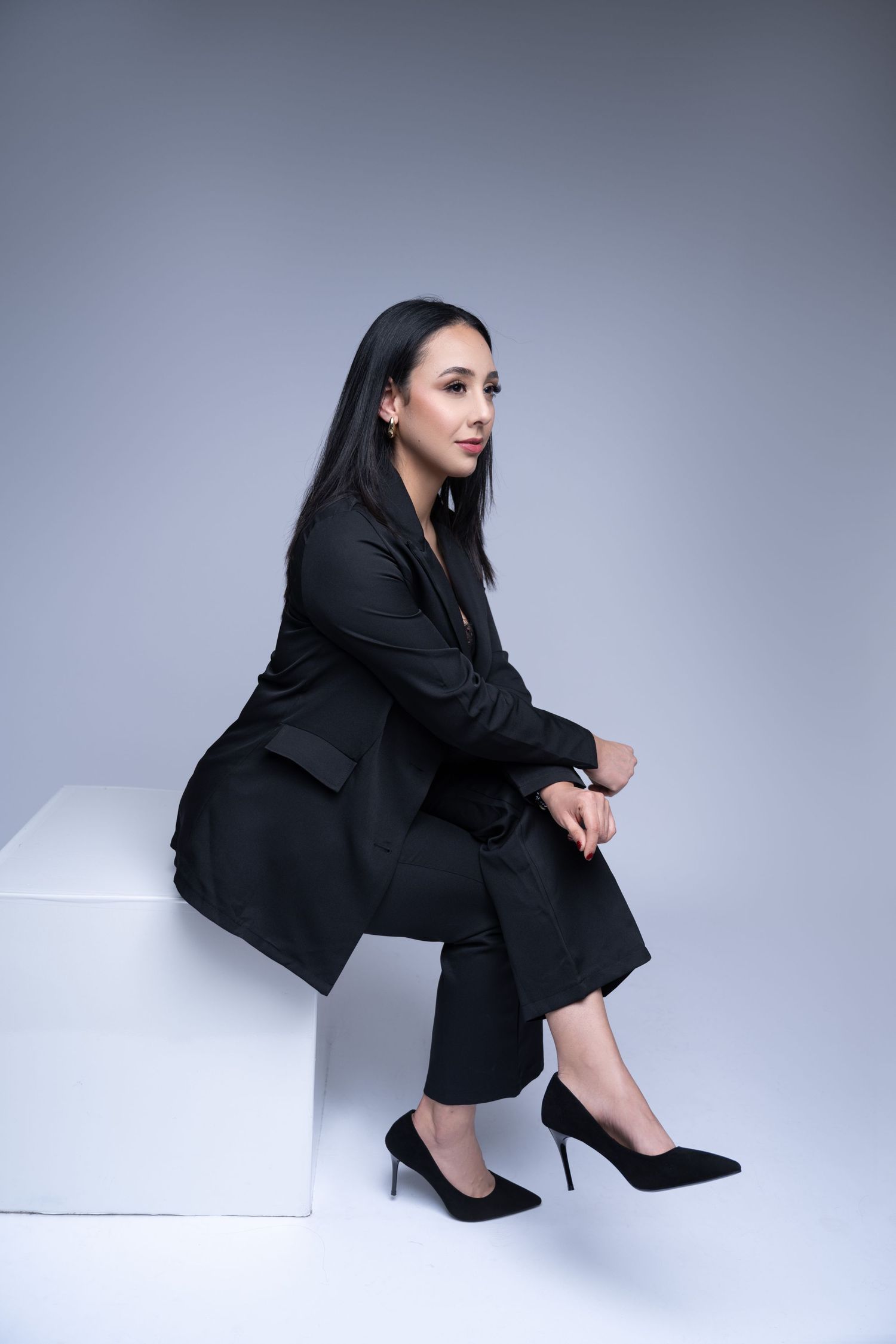 Professional woman in black business suit seated on white pedestal in studio setting.