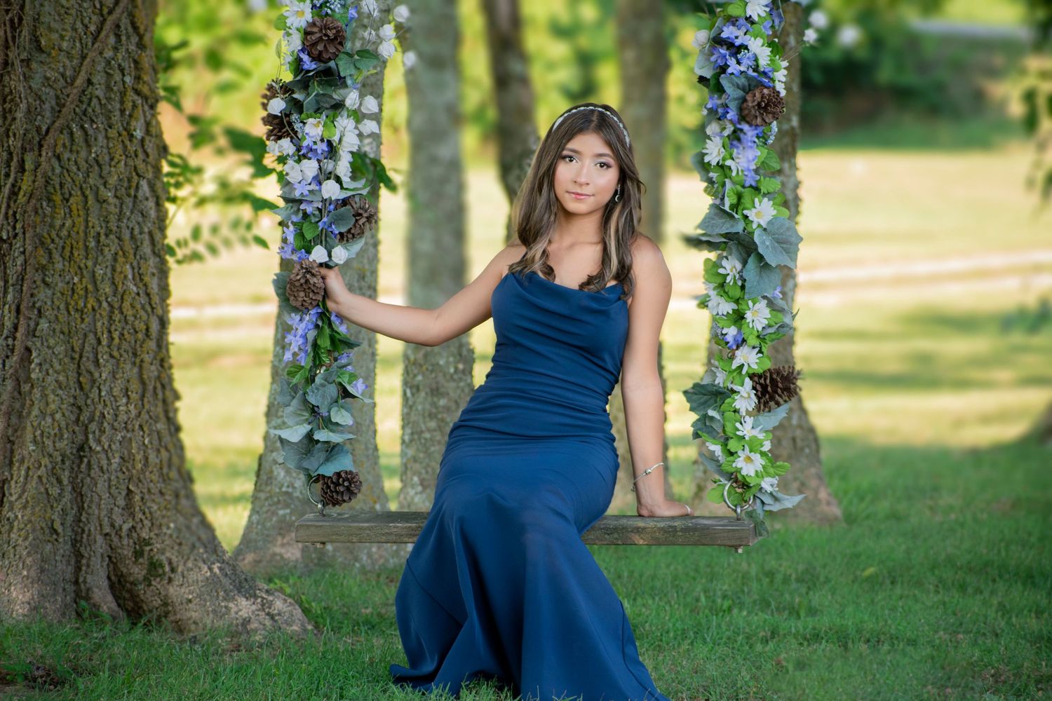 Young woman in navy blue gown posing gracefully beside a floral decorated swing between two trees outdoors.