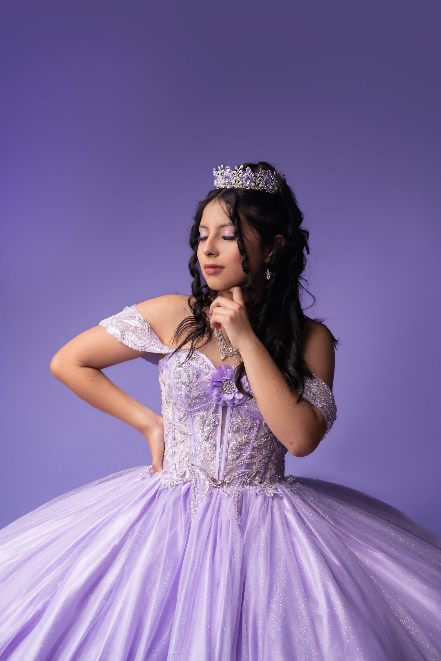 Young Latina girl in lavender quinceañera ball gown with silver tiara and lace bodice posing against purple background.