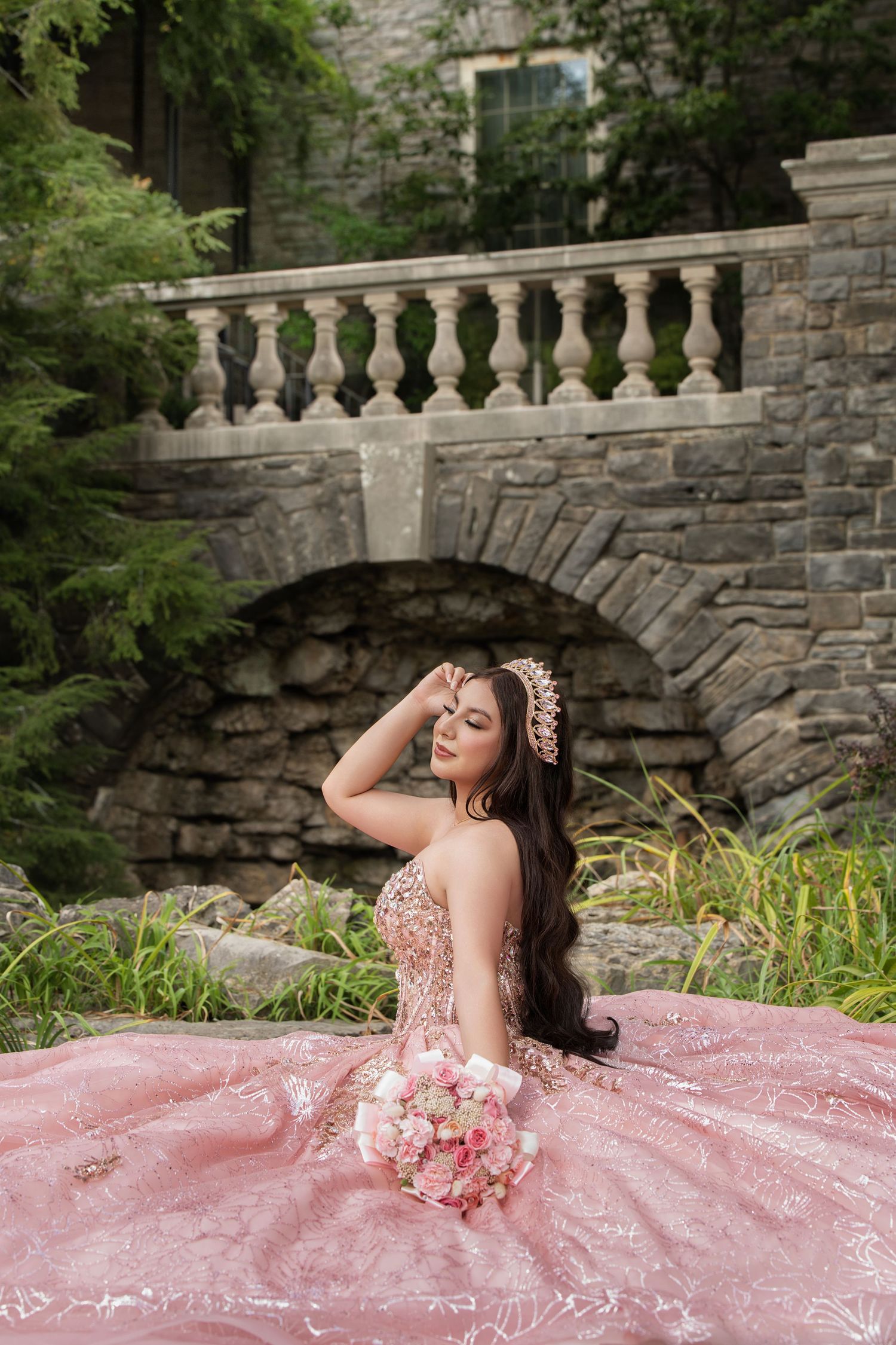 Young woman in pink quinceañera ball gown with floral crown holds bouquet before stone arch bridge.