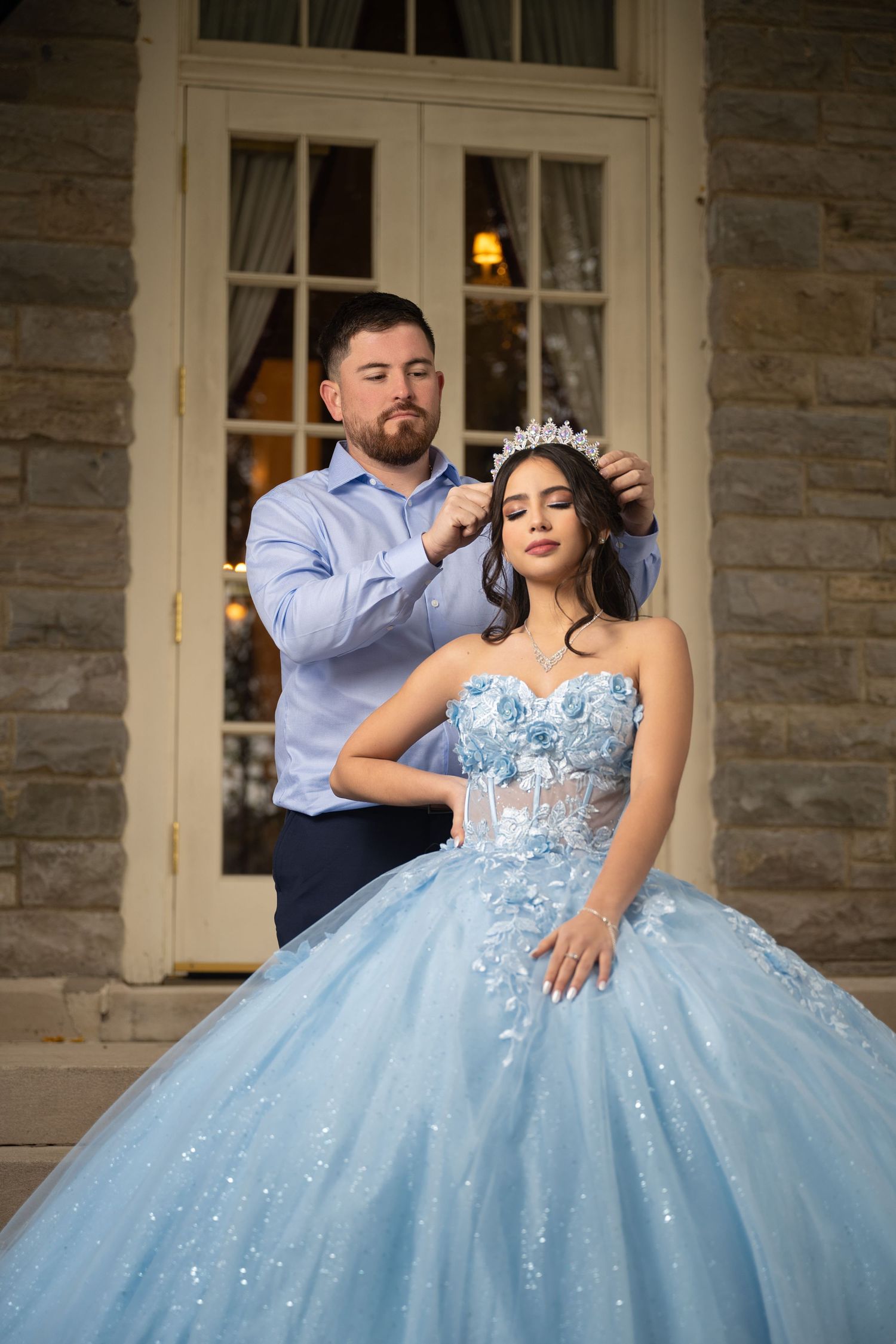 Man placing a crystal tiara on a young woman in a light blue quinceañera ball gown outside a stone building.