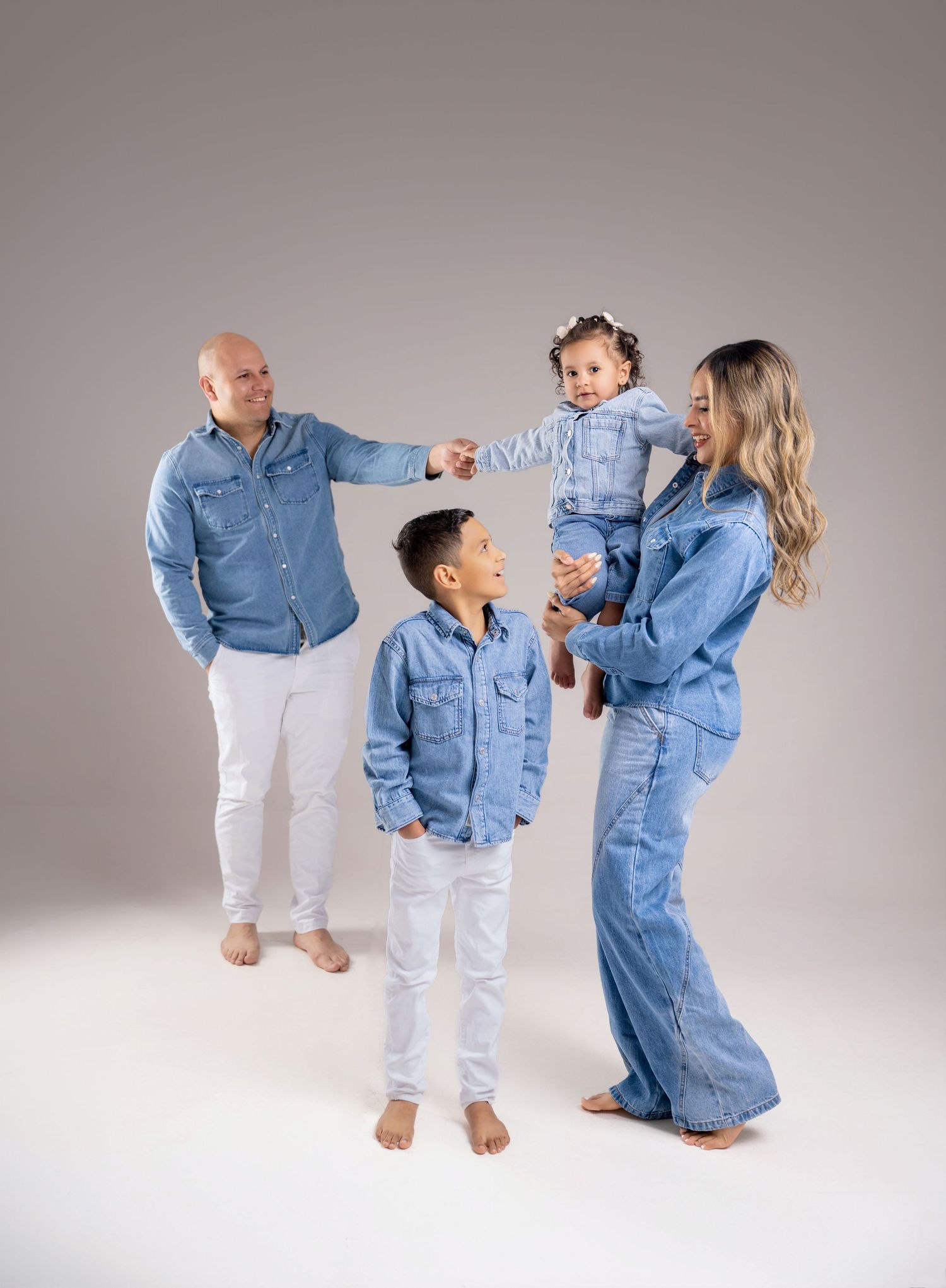 Family of four in matching denim outfits posing together in a studio, parents holding toddler while son stands smiling.