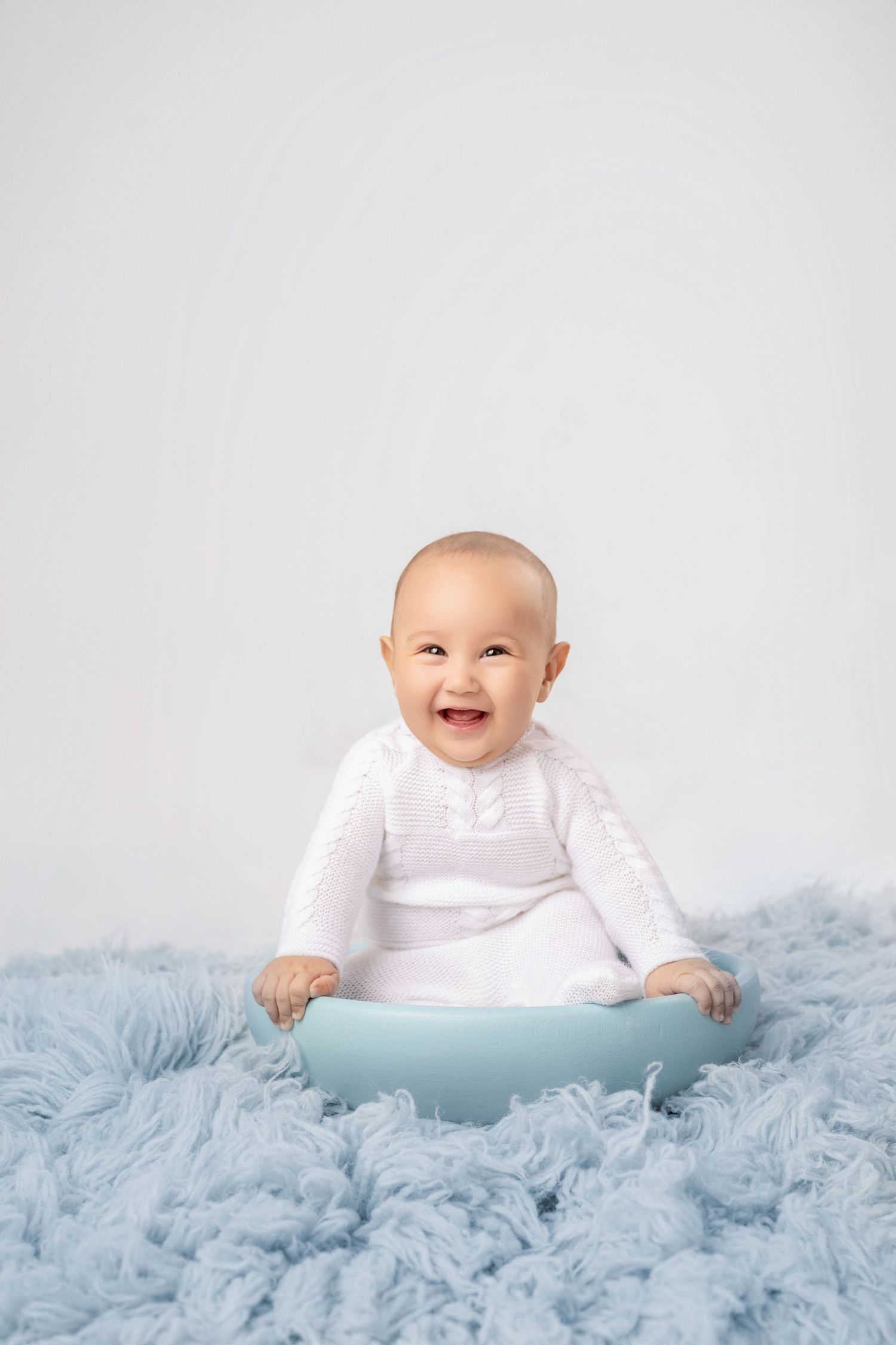 Smiling baby in white knit outfit sitting in a blue bowl on a fluffy light blue rug against a white background.