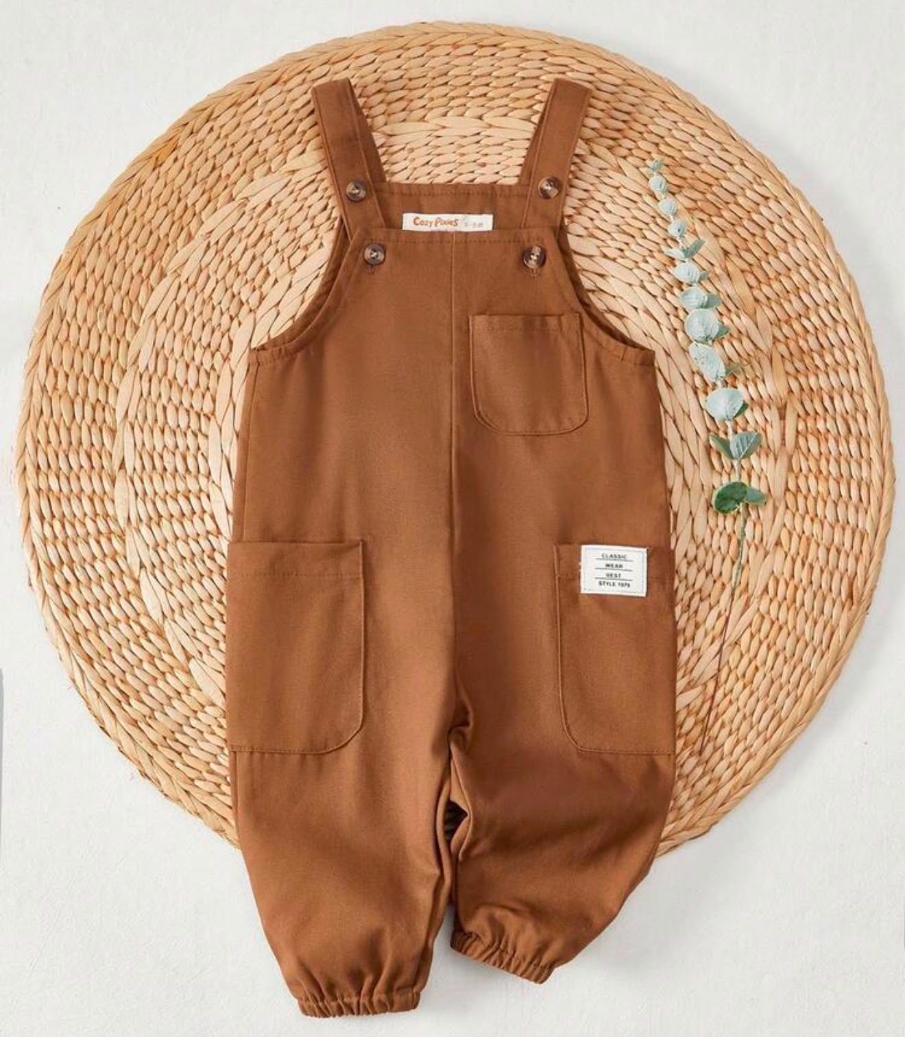 Brown cotton baby overalls with front pockets and adjustable straps, displayed on a woven rattan background.