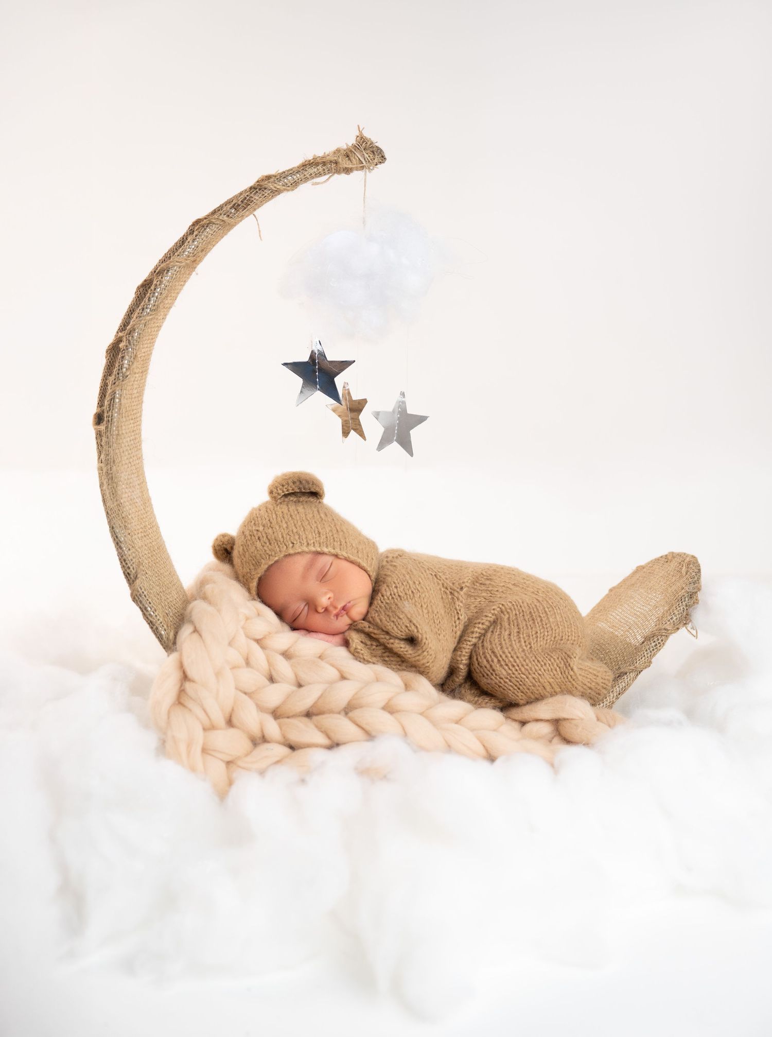 Newborn baby sleeping in a crescent moon prop wearing a bear knit outfit with star mobile decorations on white background.