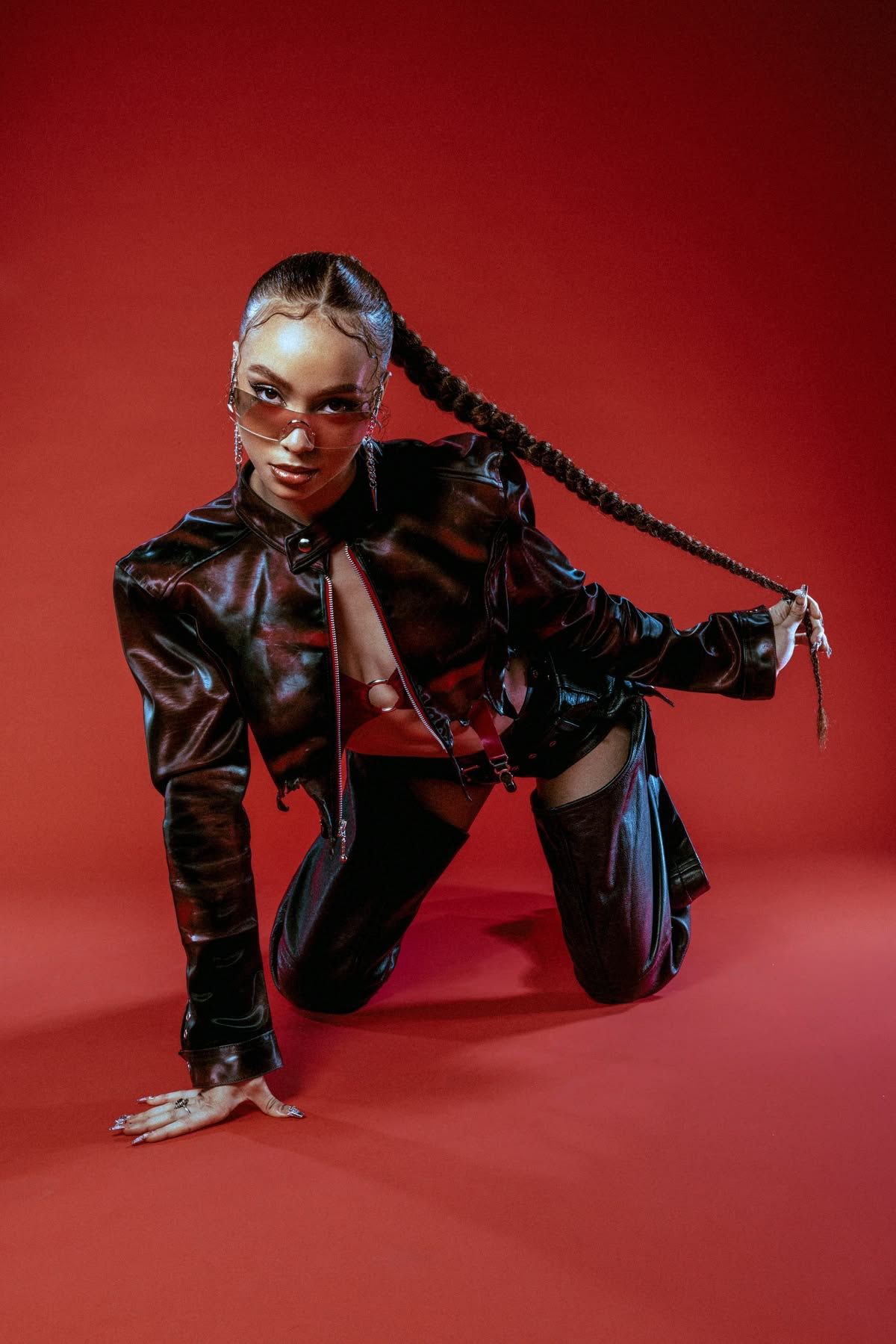 Woman in black latex outfit and braided hair poses on red background.