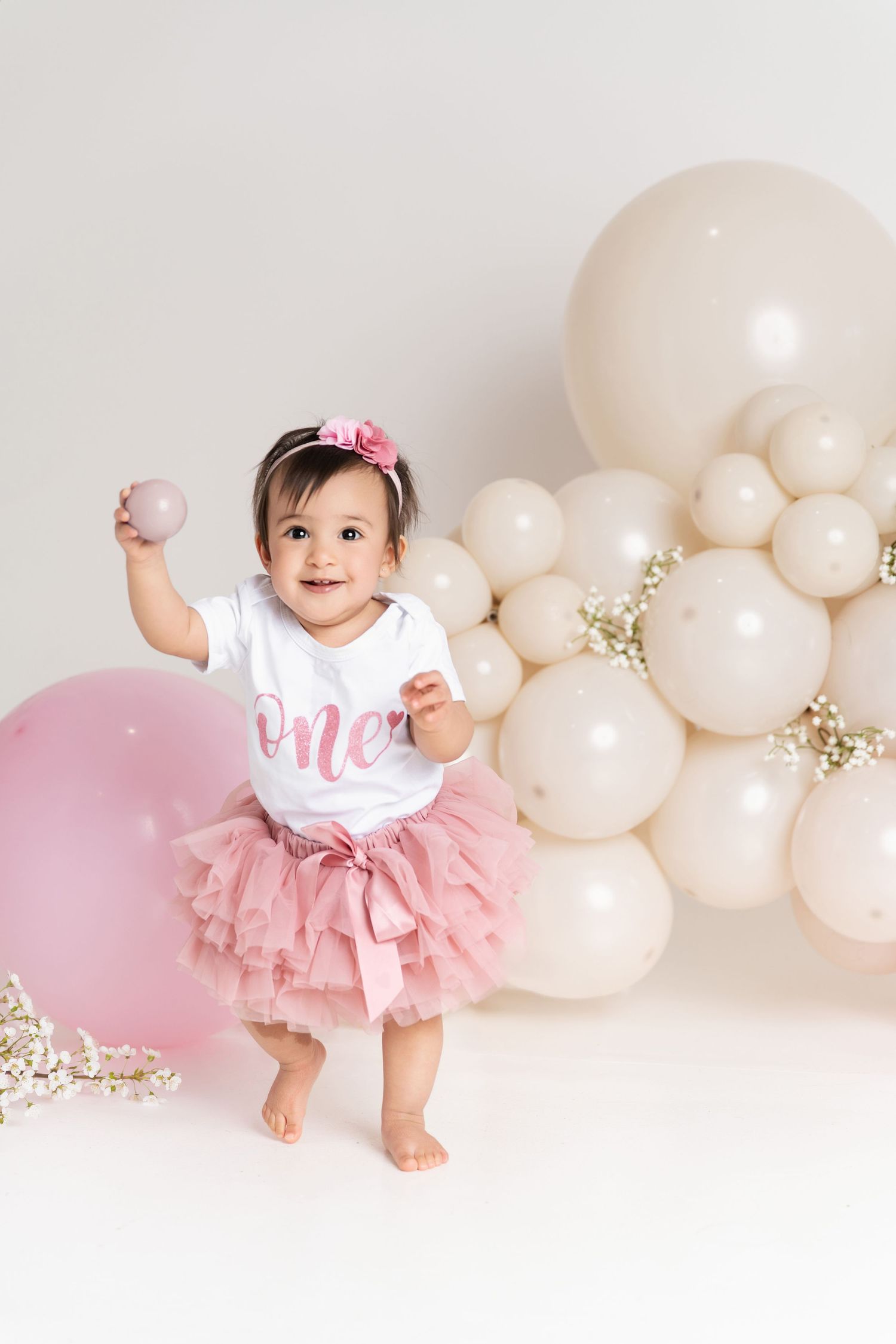 Baby girl in pink tutu and “One” onesie celebrates first birthday surrounded by cream and pink balloons.