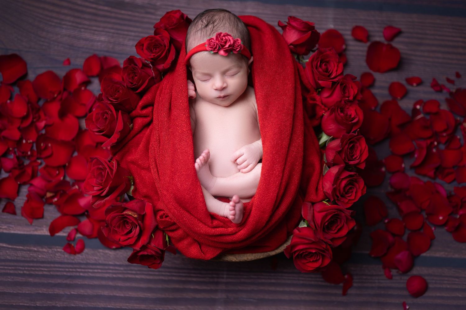 Sleeping baby nestled in red fabric surrounded by rose petals.