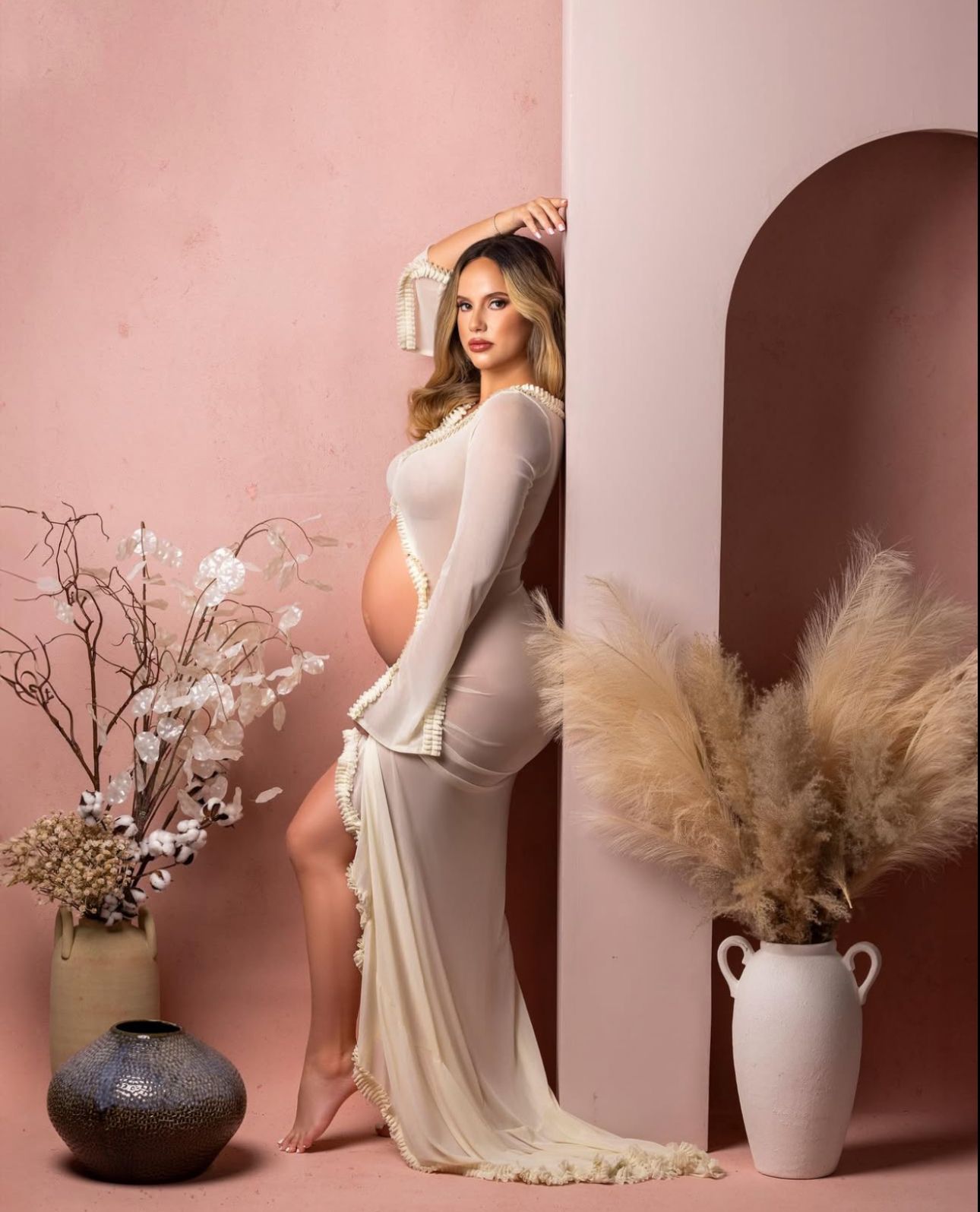 Pregnant woman in elegant cream maternity gown posing in pink studio with pampas grass and floral vases.
