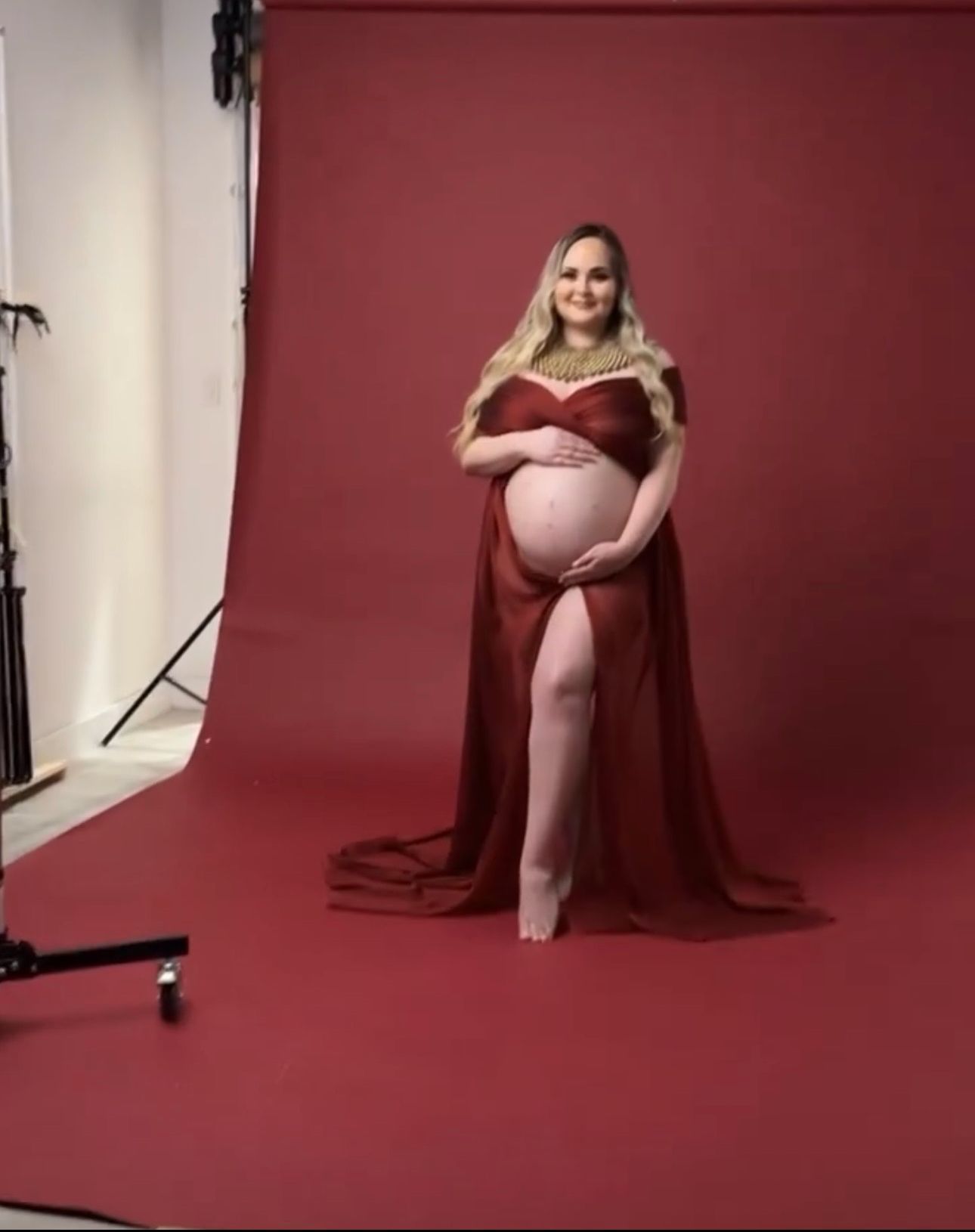 Pregnant woman in elegant red maternity gown with flowing cape poses in professional photo studio against red backdrop.