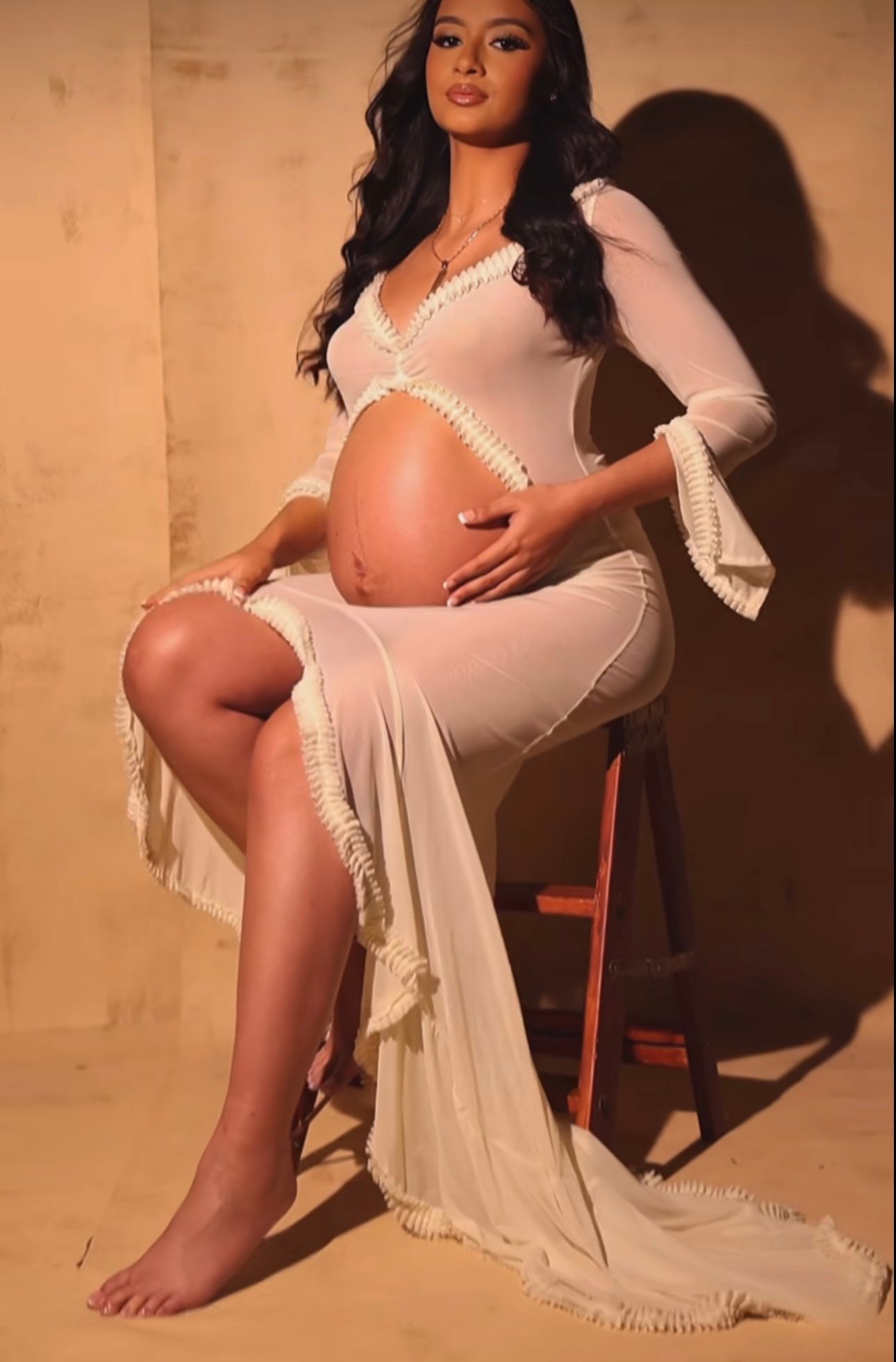 Pregnant woman in white sheer maternity gown sitting on stool, cradling baby bump during elegant maternity photoshoot.