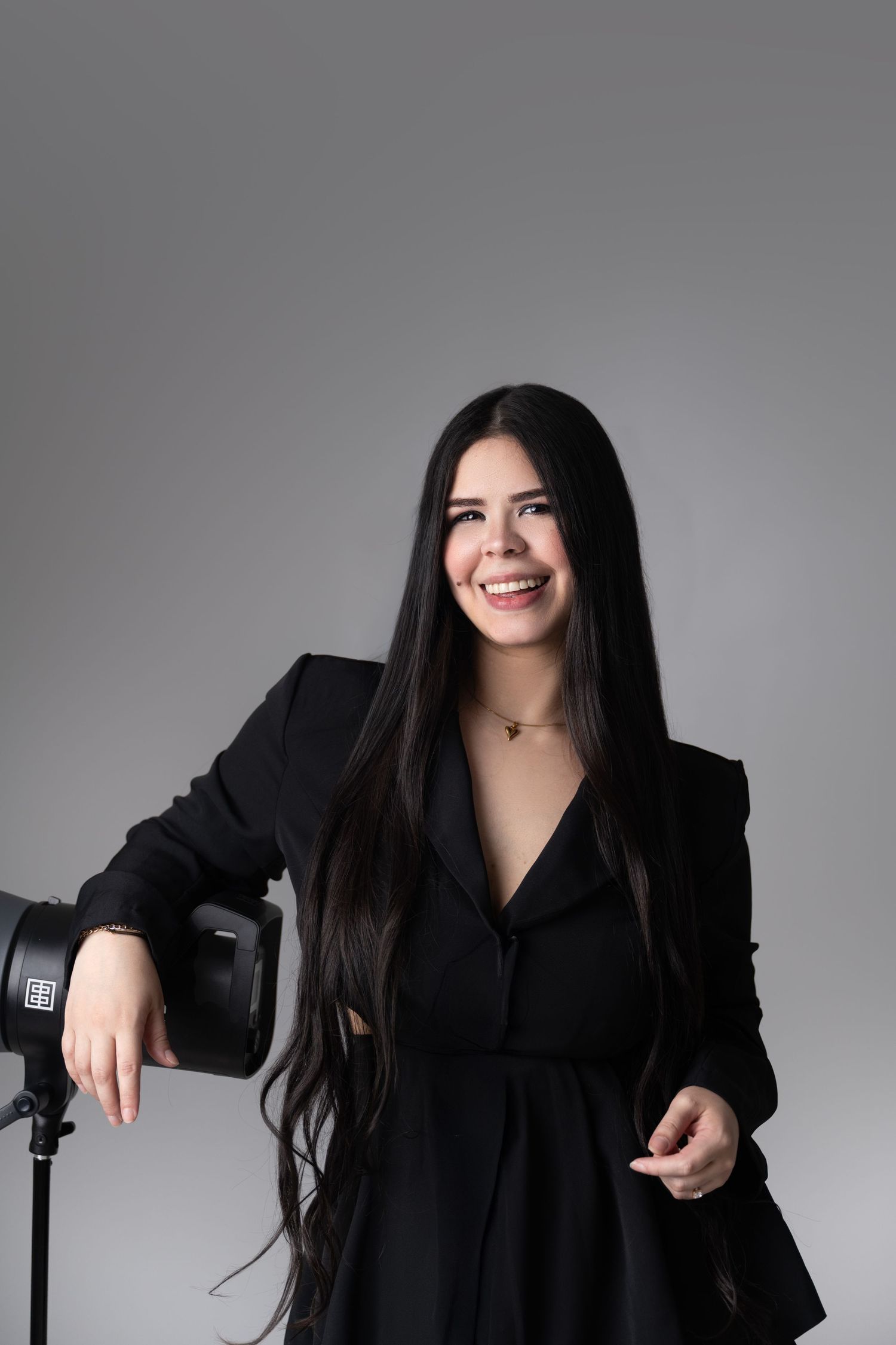 Professional studio portrait of someone in a black blazer smiling against a grey background with photography lighting equipment.