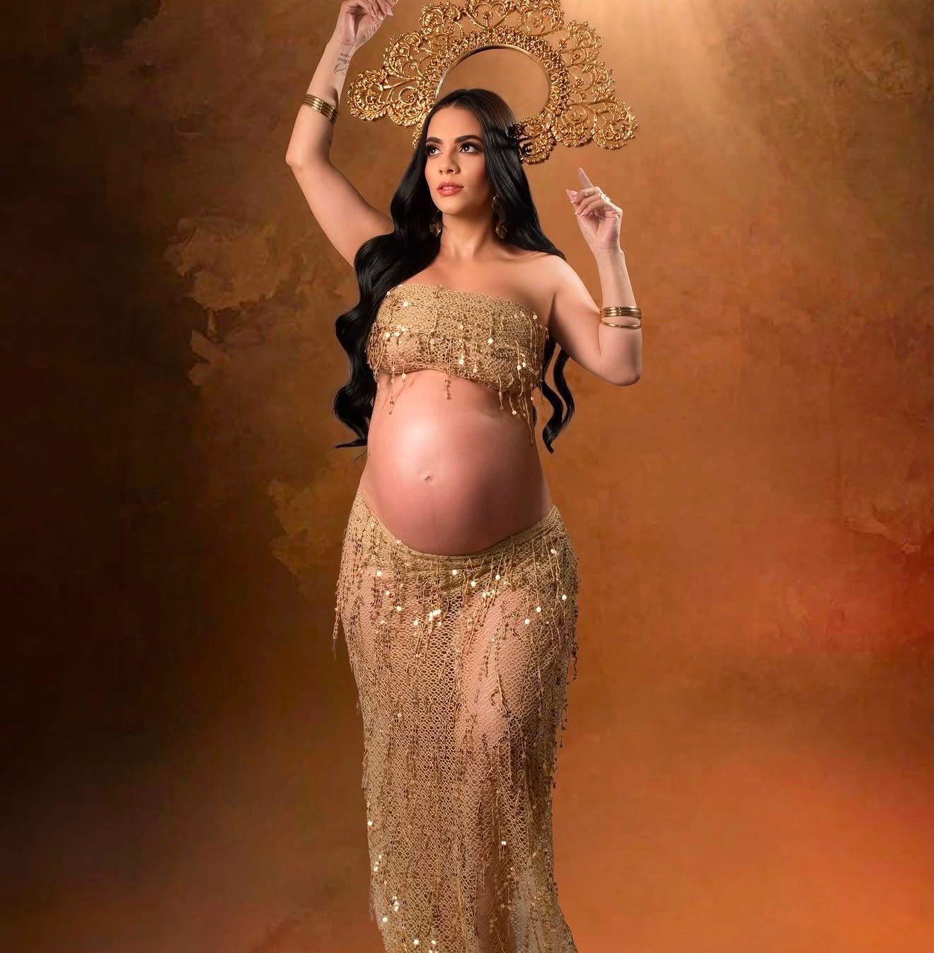 Pregnant woman in gold beaded maternity gown with ornate crown headpiece poses gracefully in studio.