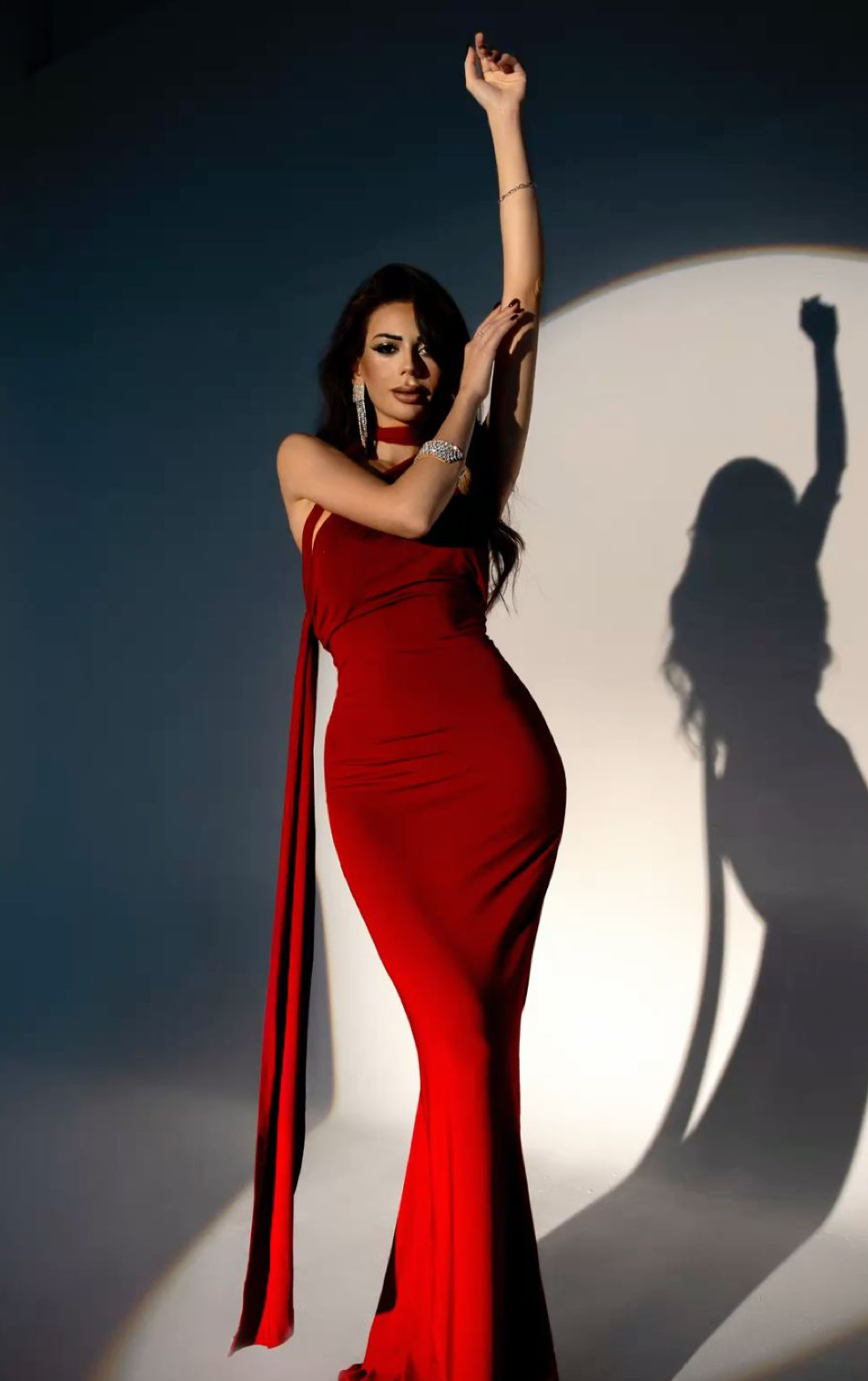 Woman in elegant red gown poses dramatically with arms raised, casting a striking shadow on a lit background.