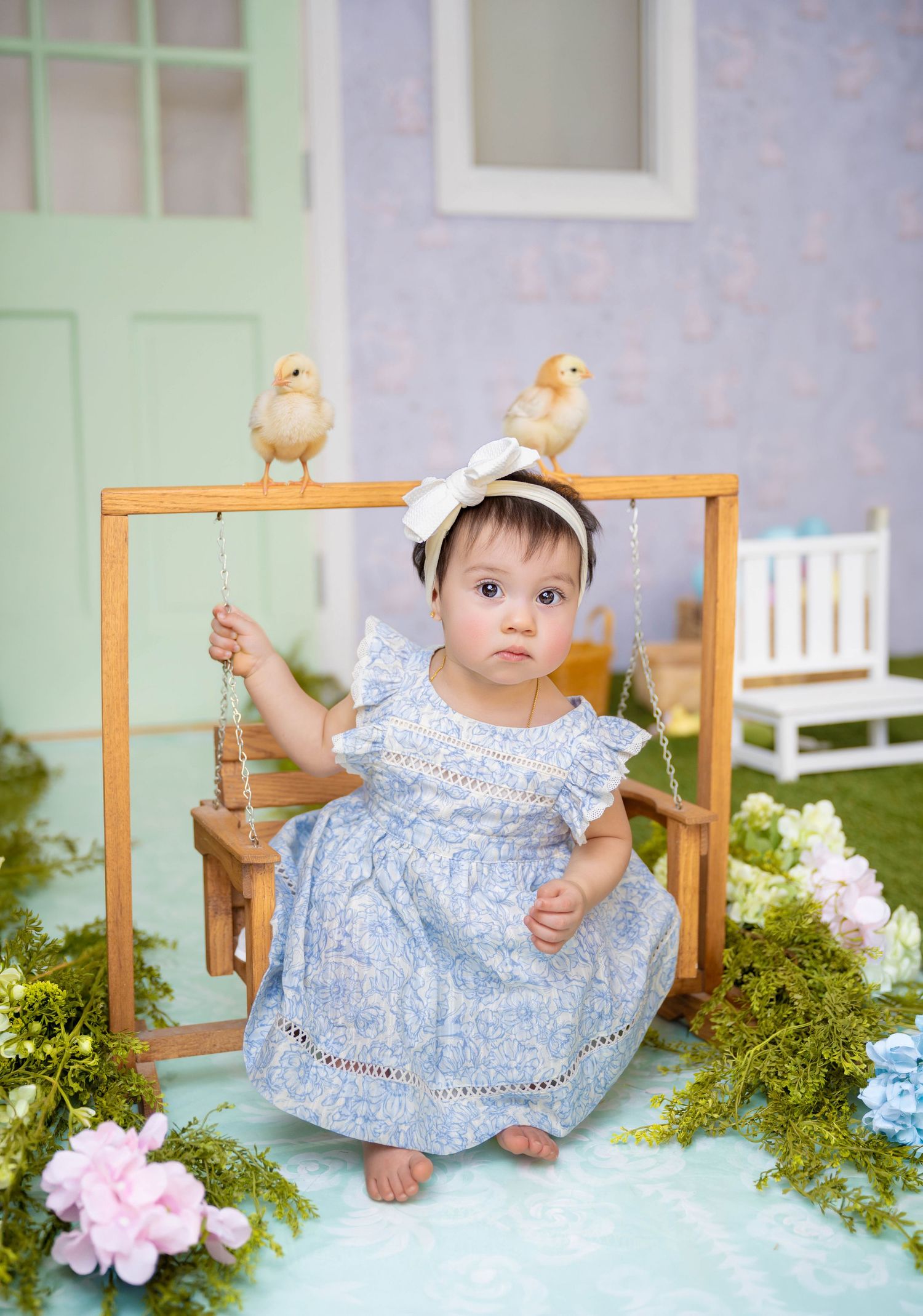 Baby girl in blue dress sits on wooden swing with chicks, surrounded by flowers in a charming spring photo setup.