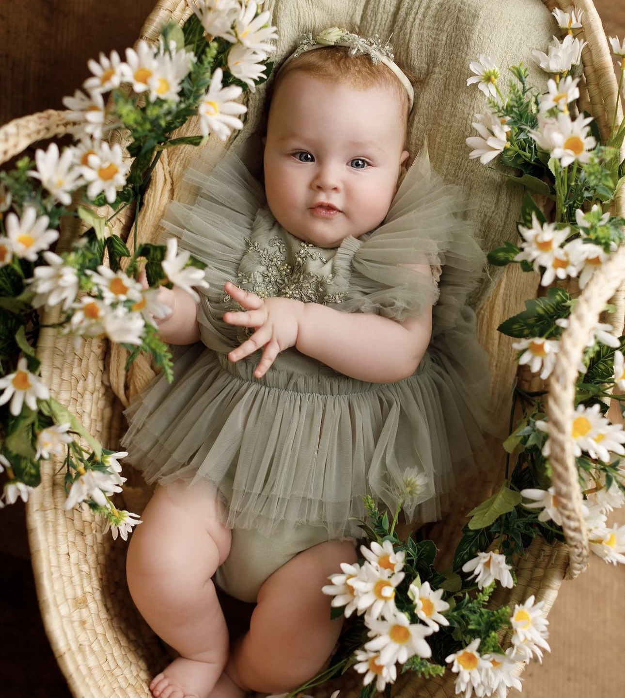 Adorable baby in sage green tulle romper lying in wicker basket surrounded by white daisy flowers.