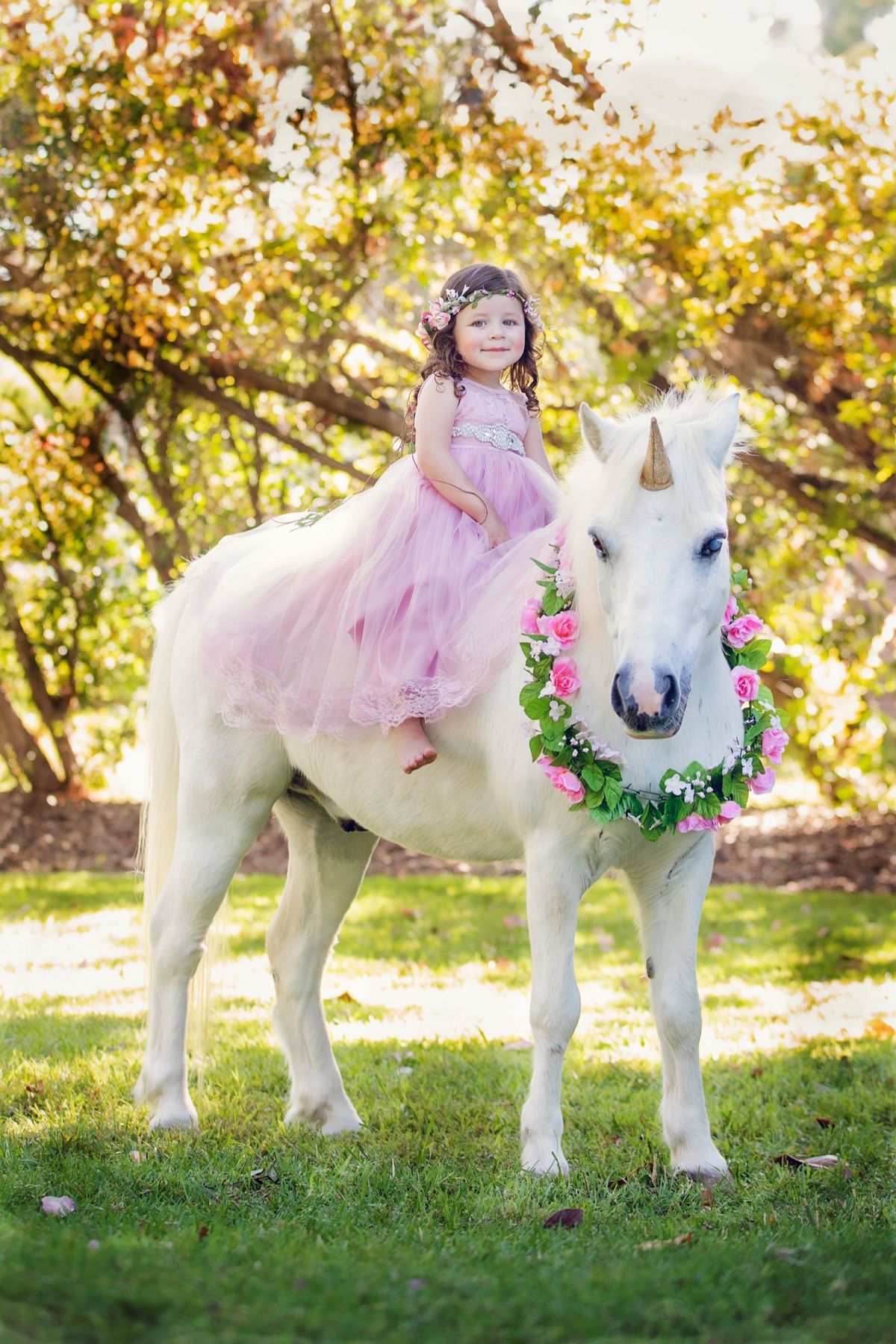 Young girl in pink tulle dress sitting on a white unicorn with floral wreath in a sunny garden.