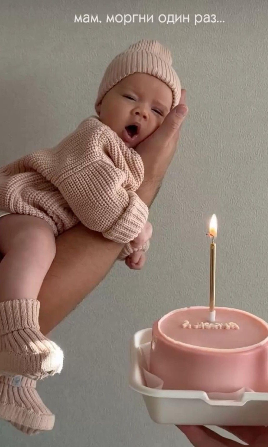 Sleeping newborn baby in pink knit outfit held near a pink birthday cake with one lit candle.