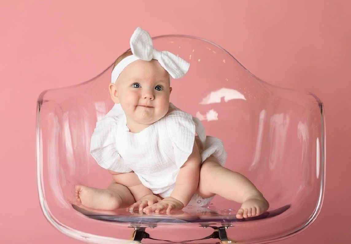 A baby wearing a white bow lies in a clear glass bowl against a pink background.