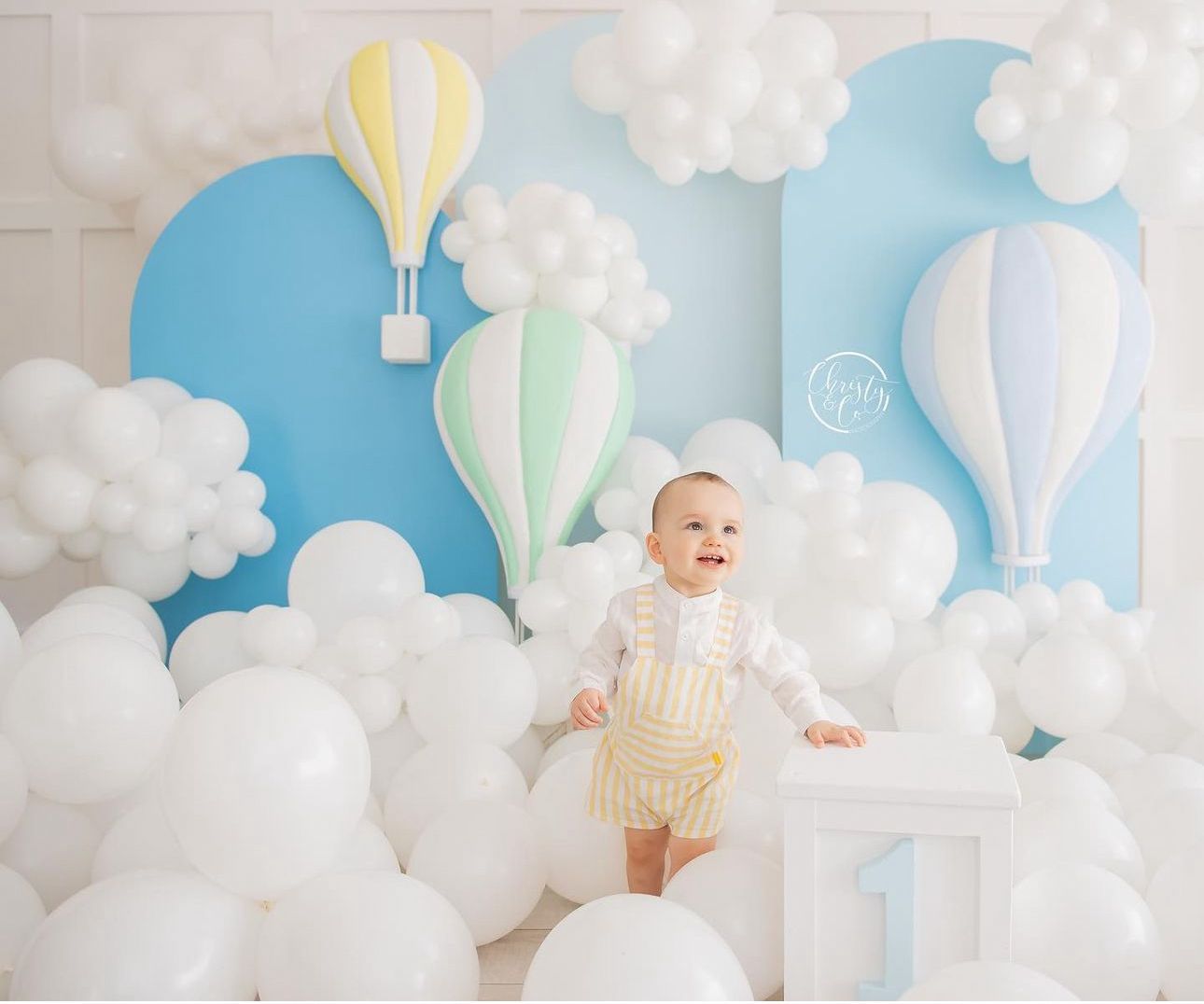A baby in a white outfit sits among pastel hot air balloon decorations and fluffy white clouds in a dreamy nursery setting.
