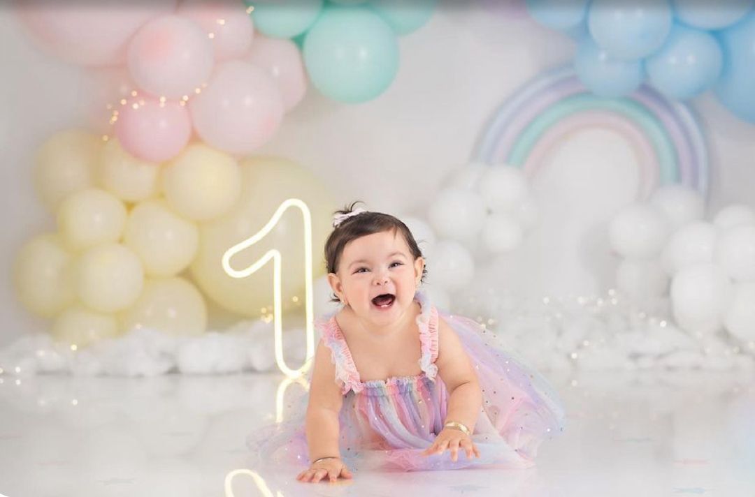 Baby in pastel pink and blue tulle dress crawls on white floor with soft colored balloons in background.