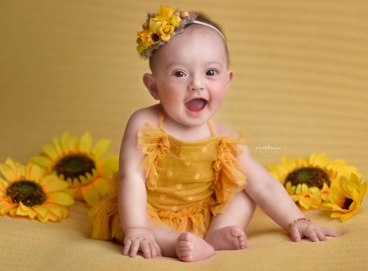 Smiling baby in yellow outfit sits on yellow backdrop with decorative sunflowers.
