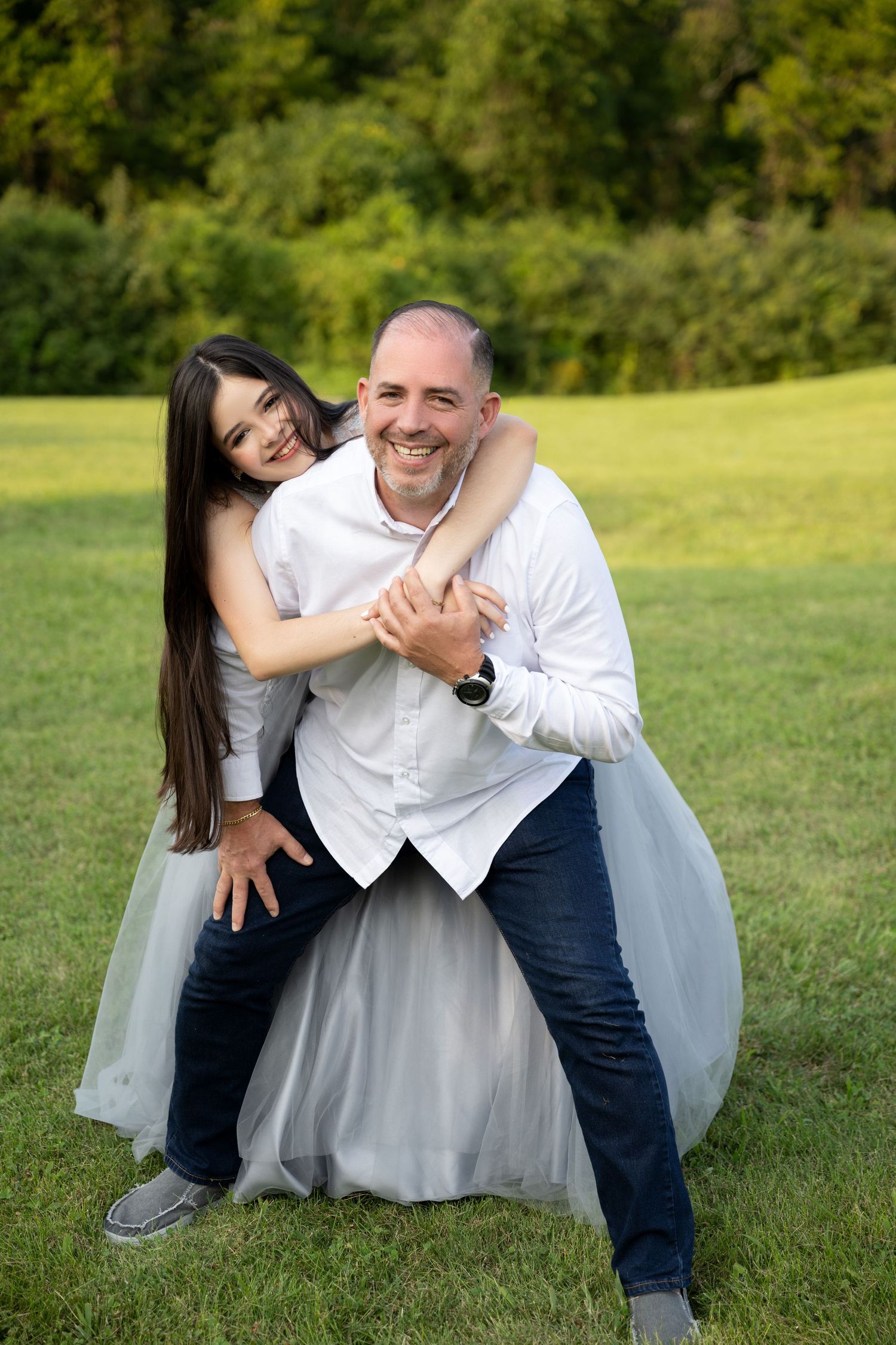 A smiling couple poses outdoors on green grass, woman in white tulle dress hugging man in white shirt from behind.