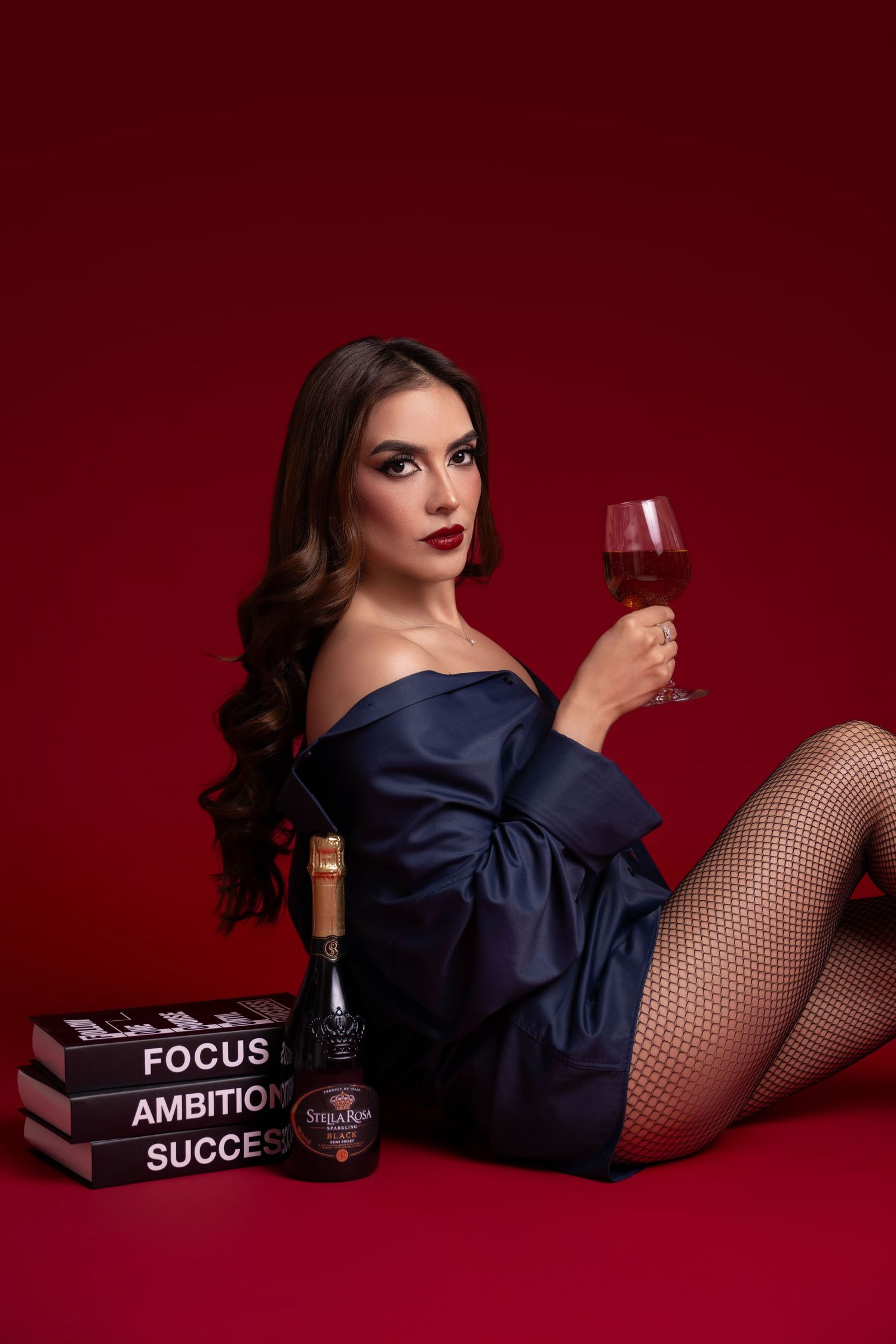 Woman in an off-shoulder navy shirt and fishnet tights holds a glass of red wine beside a wine bottle and books on a red backdrop.