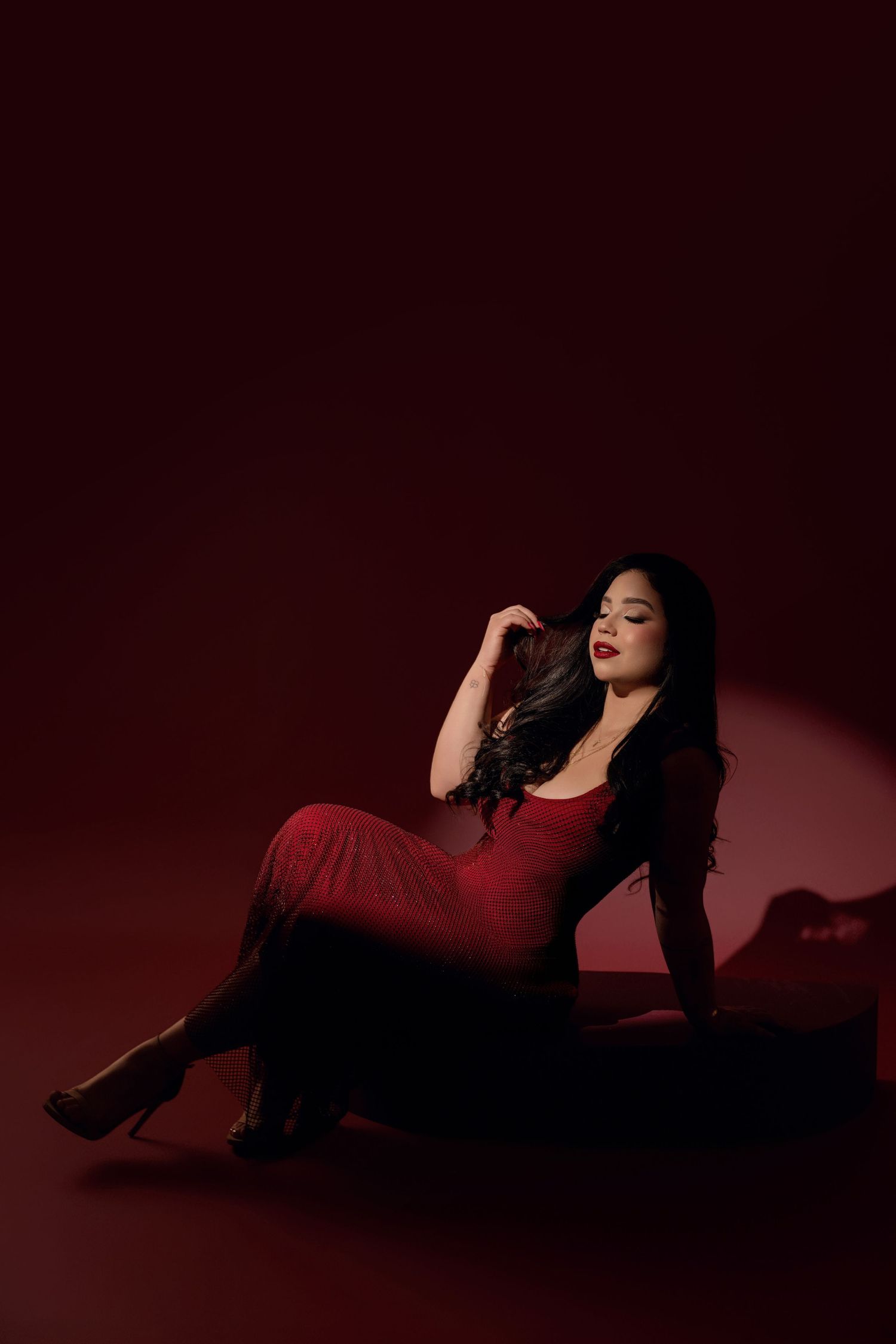 Woman with long dark wavy hair and red lipstick poses seated in a red dress and heels against a deep red studio backdrop.