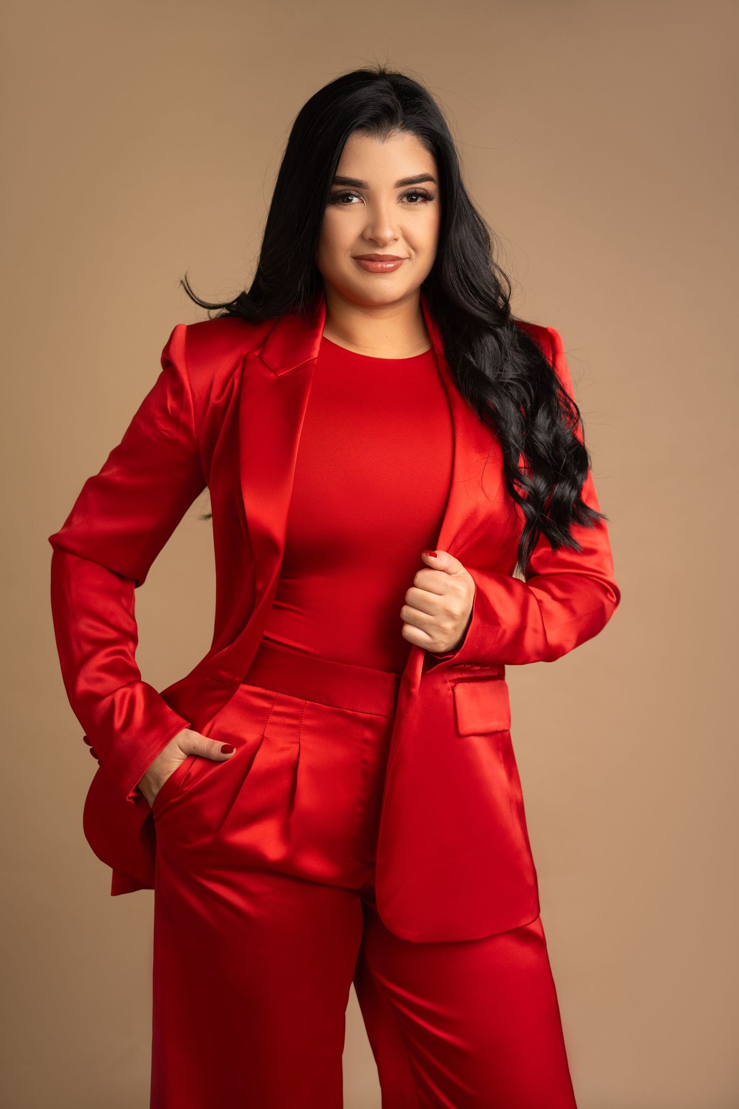 Woman in red satin suit jacket and pants posing confidently against a beige background.
