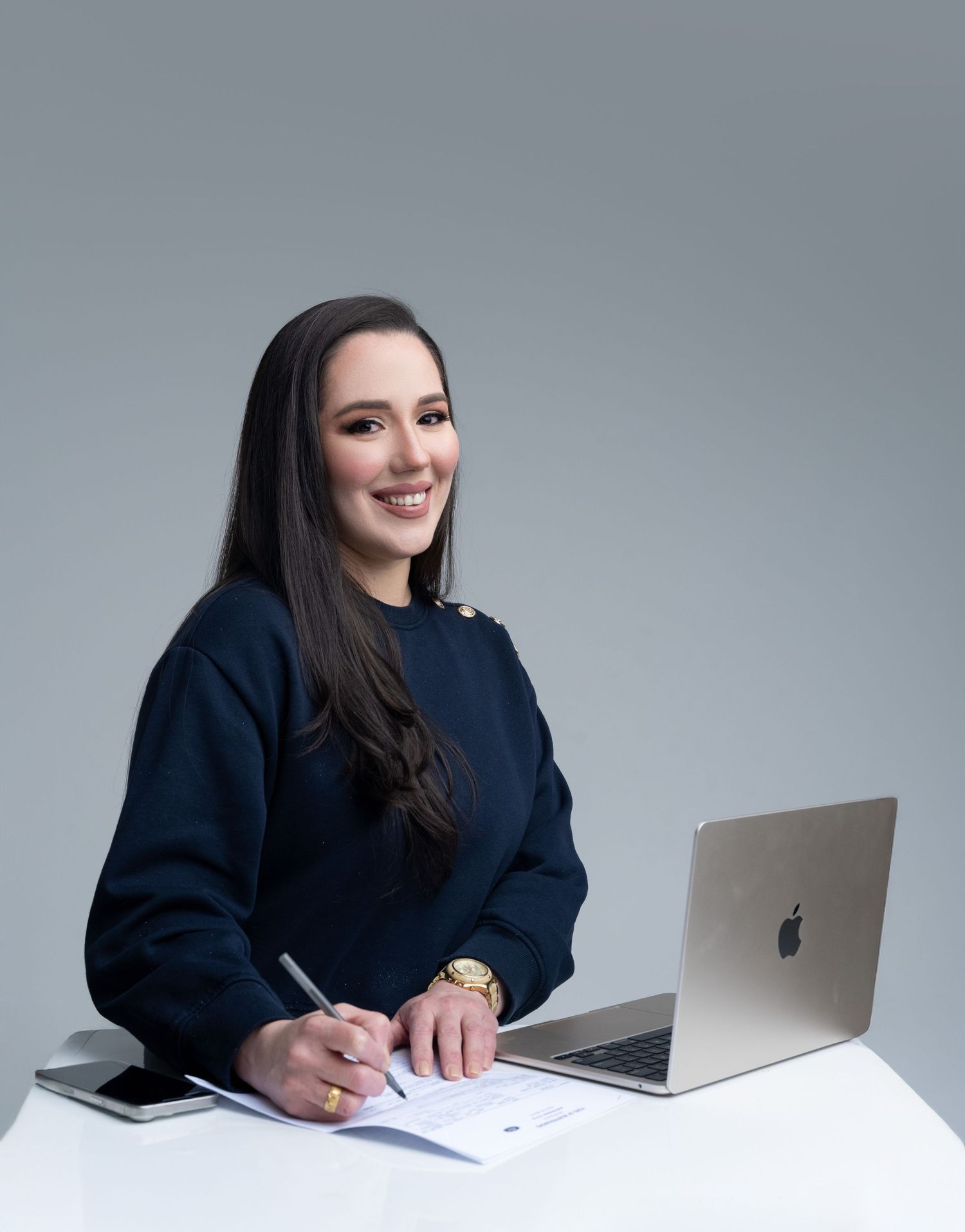 A smiling professional woman in a navy sweater sits at a white desk with an Apple laptop, writing notes.