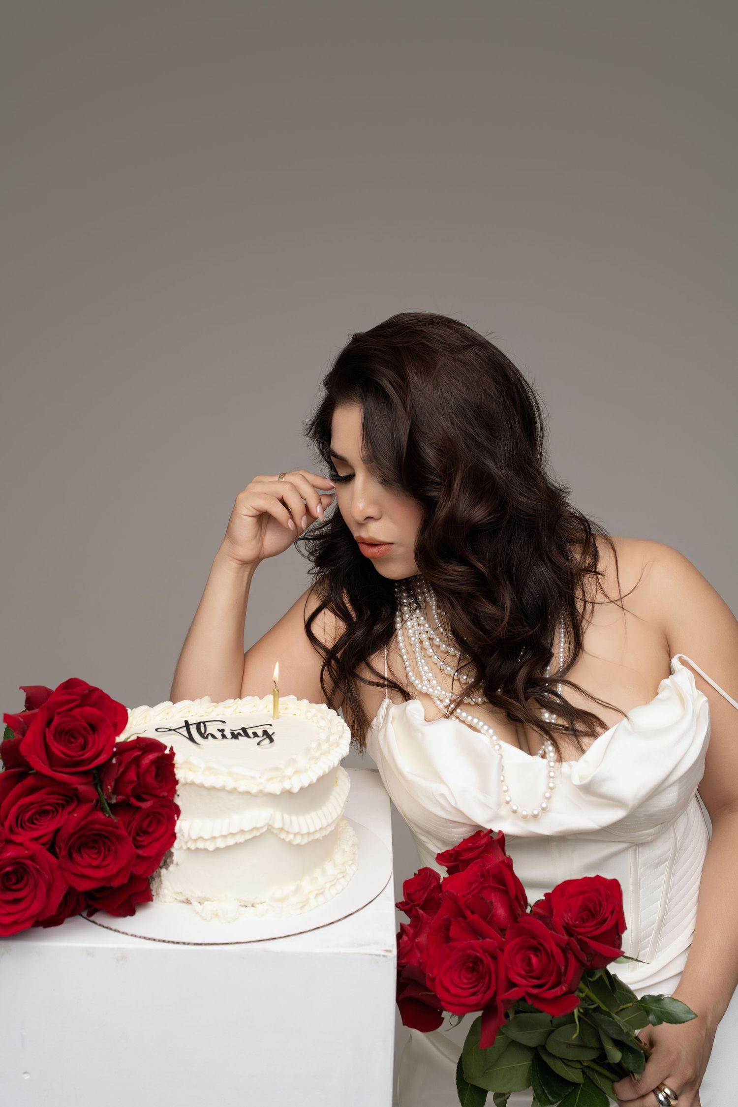 Woman in white dress gazes at a two-tier birthday cake reading thirty, surrounded by red roses on a gray background.