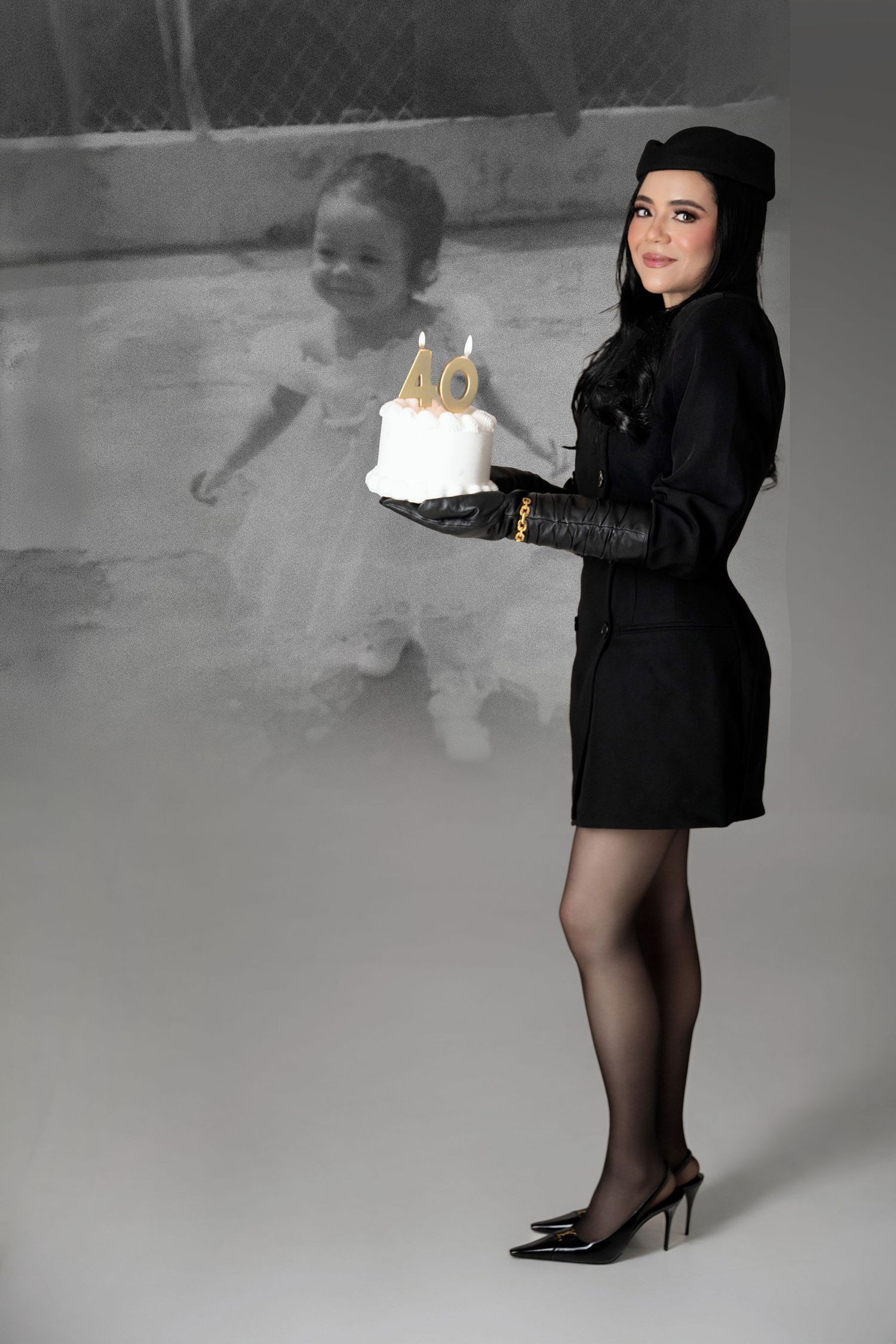 Woman in a black coat, hat, gloves and heels holds a white 40th birthday cake beside a faded childhood photo of herself in a tutu.