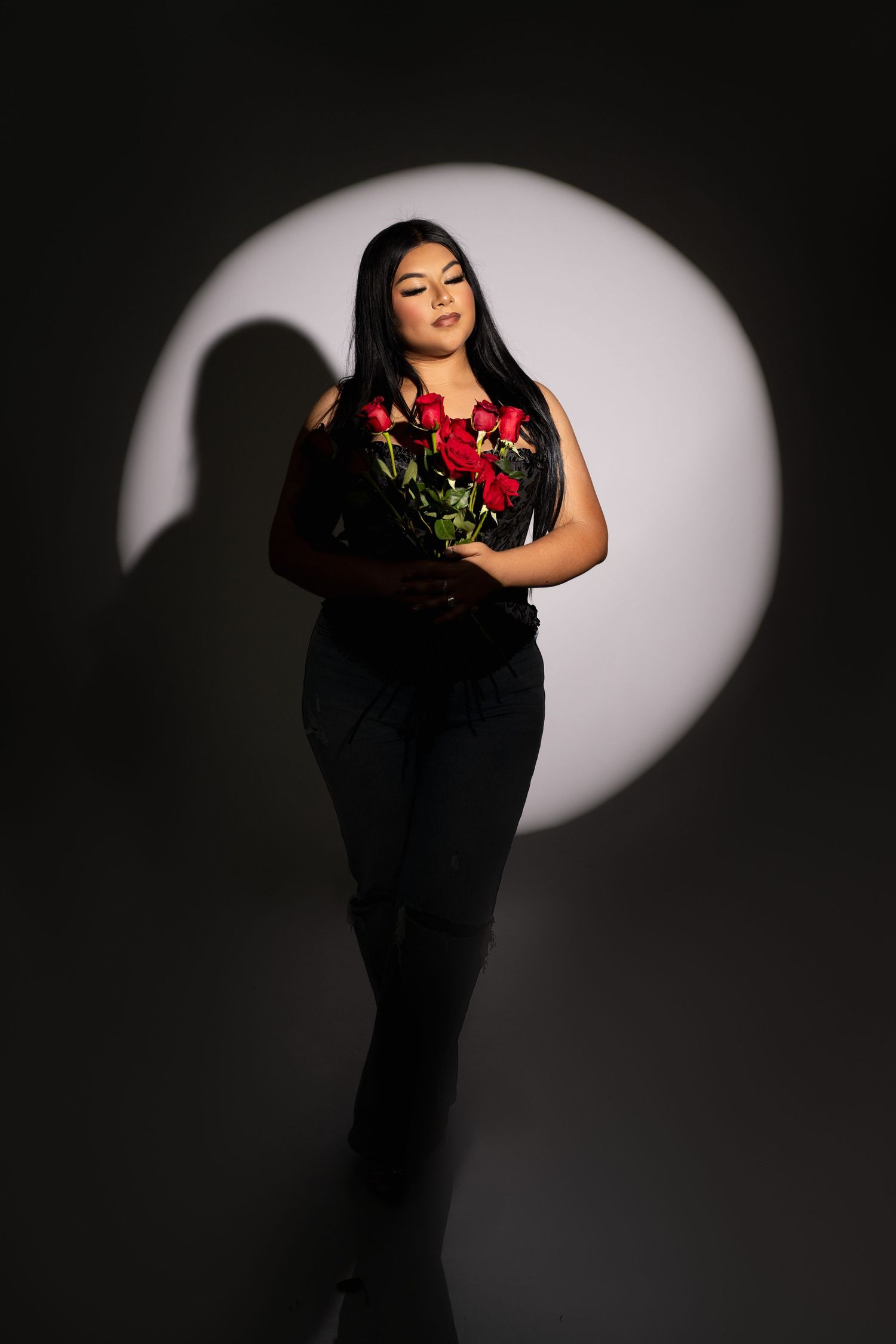 Woman with long dark hair holds a bouquet of red roses while standing in a circular spotlight against a dark studio backdrop.