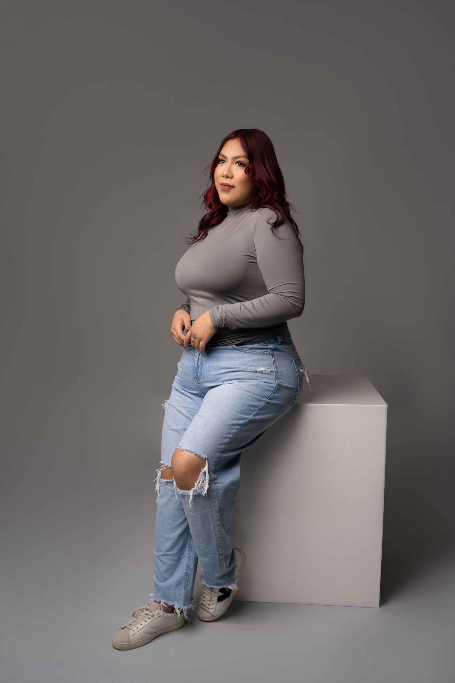 Woman with long auburn hair, gray turtleneck and ripped light-wash jeans, seated on a white cube in a studio with a gray backdrop.