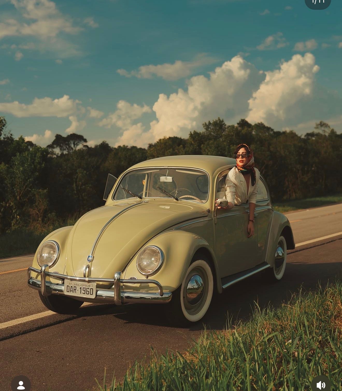 Person leaning out of a classic yellow Volkswagen Beetle on a scenic road under a cloudy blue sky.