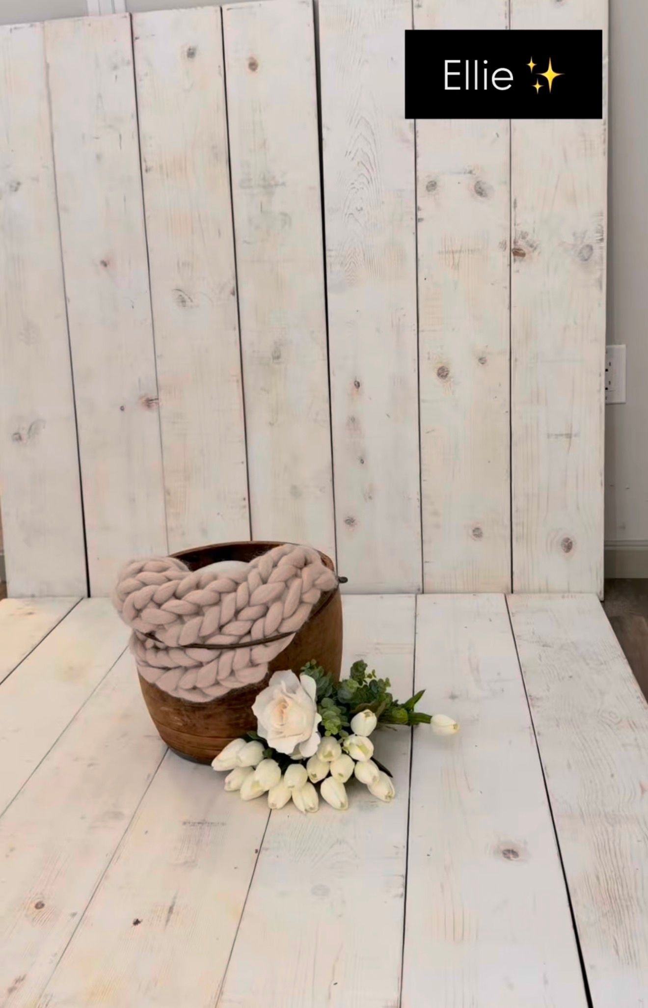 A wooden bowl with chunky knit blanket and white flowers on a whitewashed wood floor backdrop labeled Ellie.