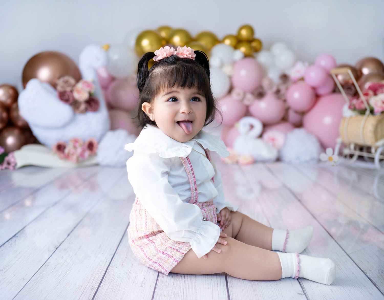 Child in white and pink outfit sits on bench with balloon and floral decorations.